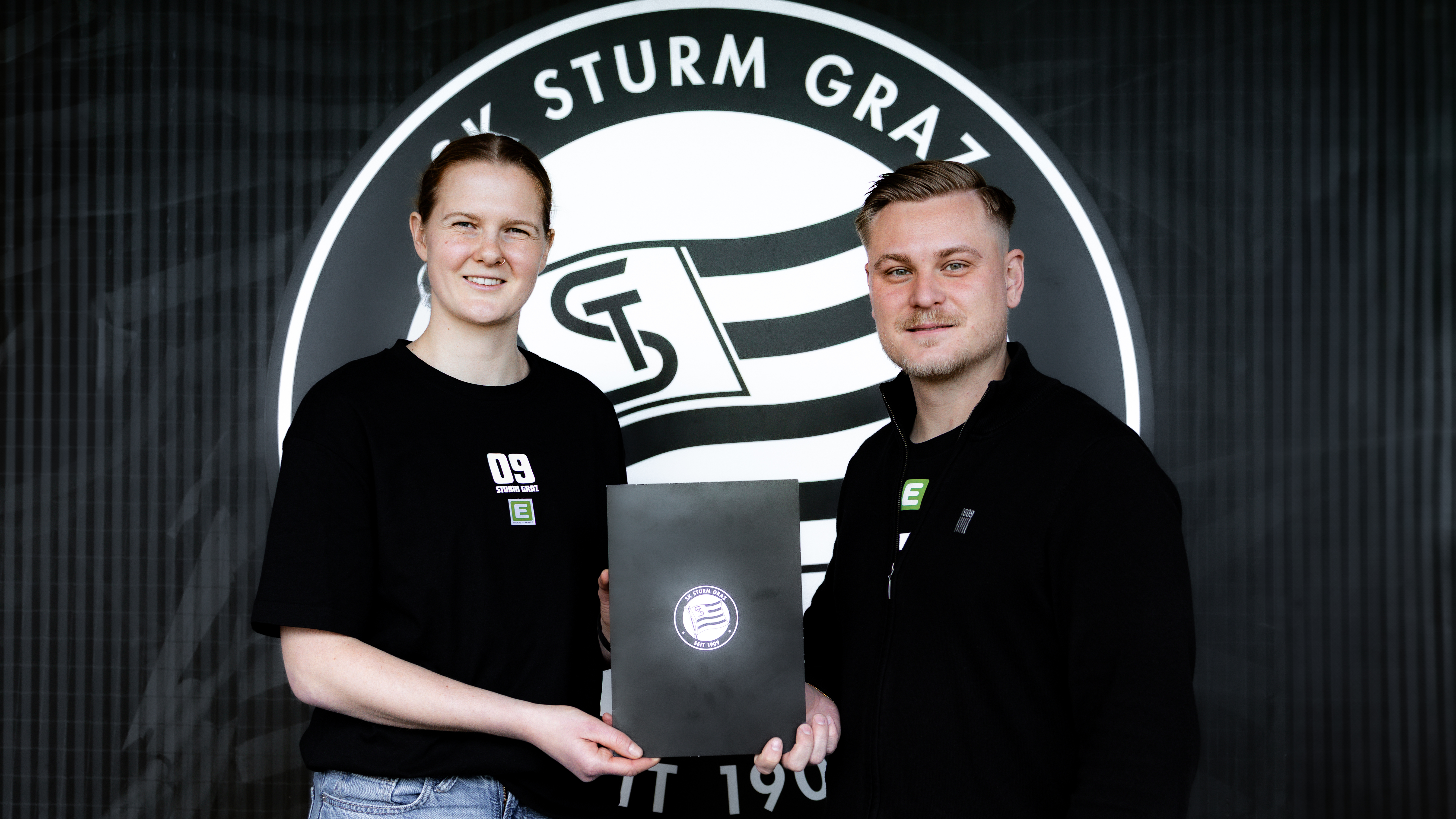 Ein Mann und eine Frau stehen vor einem großen Logo und halten einen Block hoch.