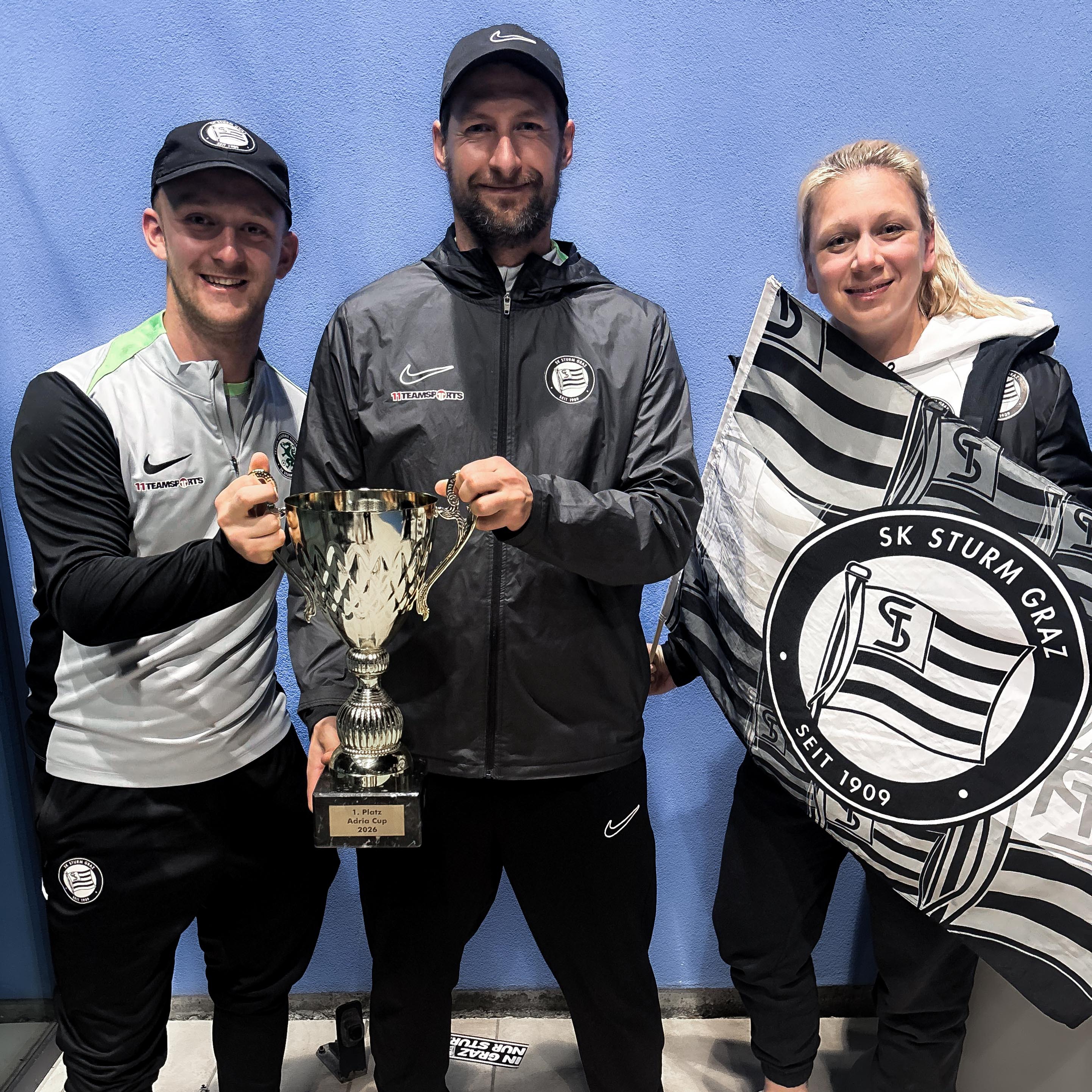 Zwei Männer halten einen Pokal und eine Frau eine Fahne