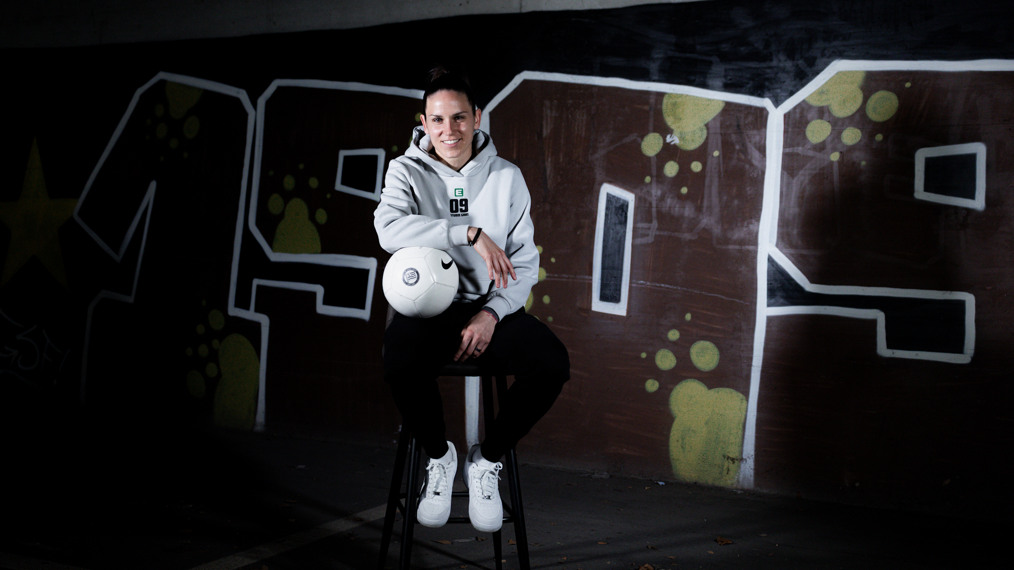 Eine Spielerin in einem Hoodie sitzt auf einem Stuhl, hält einen Ball und im Hintergrund ist ein Graffiti zu sehen.