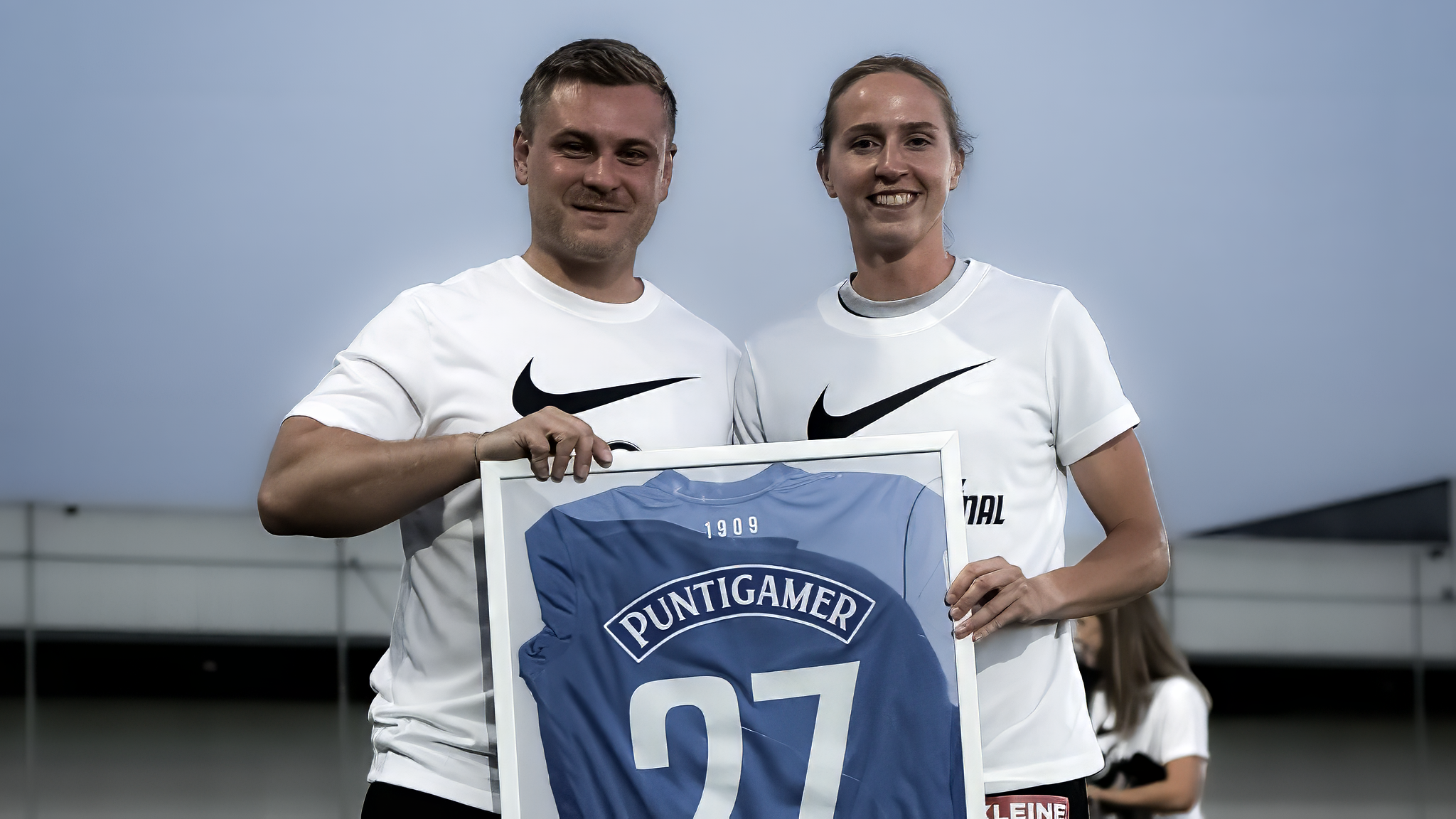 Ein Mann und eine Frau beide in einem weißen Shirt halten einen Bilderrahmen wo ein blaues Trikot zu sehen ist.