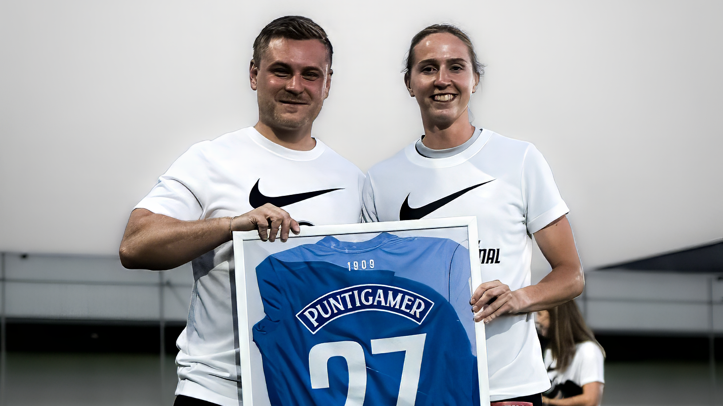 Ein Mann und eine Frau in weißen Shirts halten ein blaues Trikot in einem Rahmen hoch.