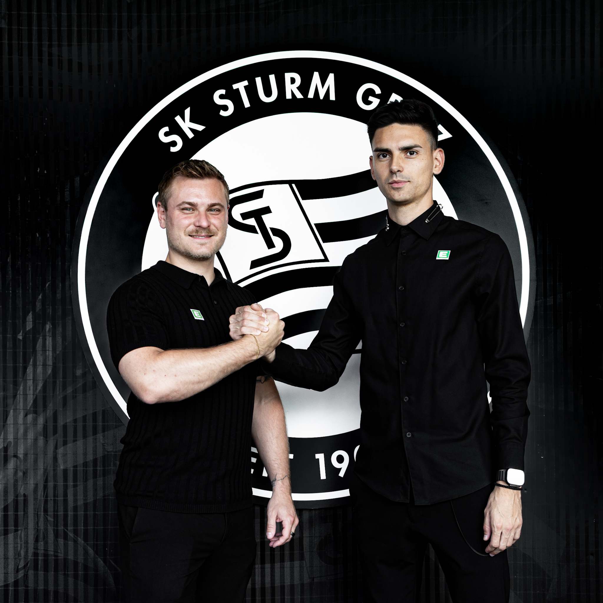Zwei Männer mit schwarzen Oberteilen geben sich die Hand vor einem schwarz-weißen Logo