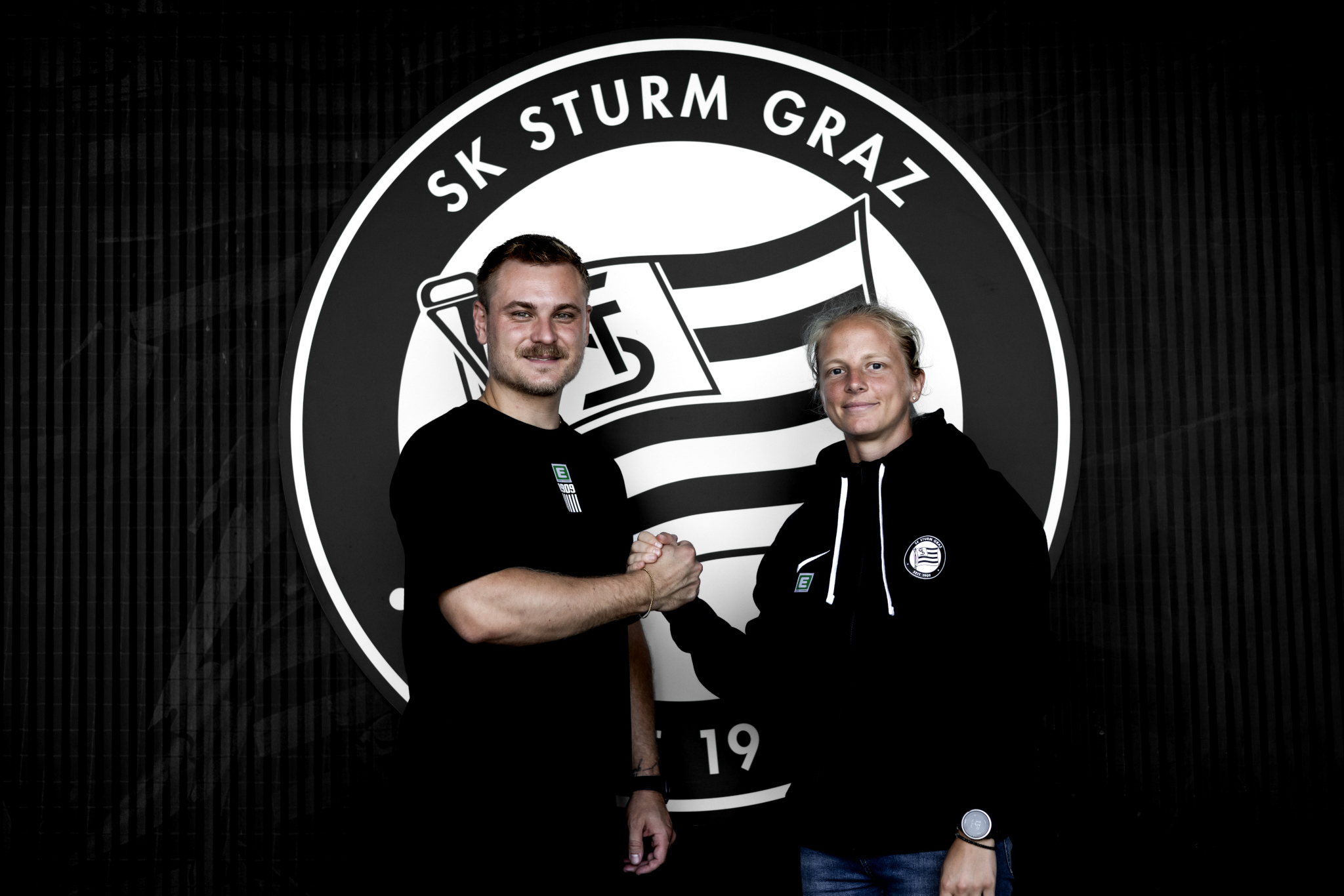 Ein Mann und eine Frau mit schwarzen Shirts geben sich vor einem schwarz-weißen Logo die Hand
