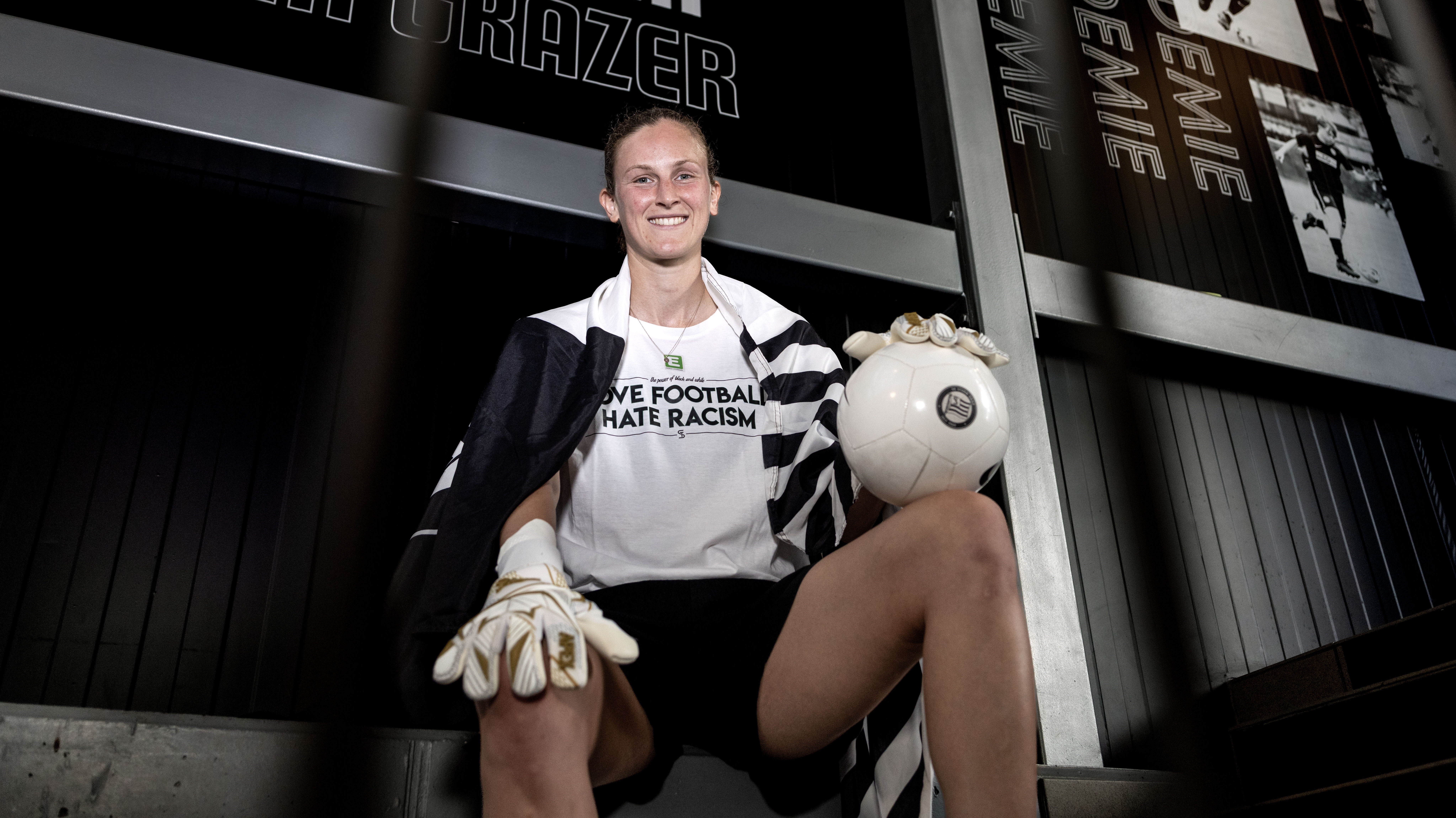 Eine Frau in einem weißen Shirt sitzt auf einer Stiege und hält mit Tormannhandschuhen einen weißen Ball.