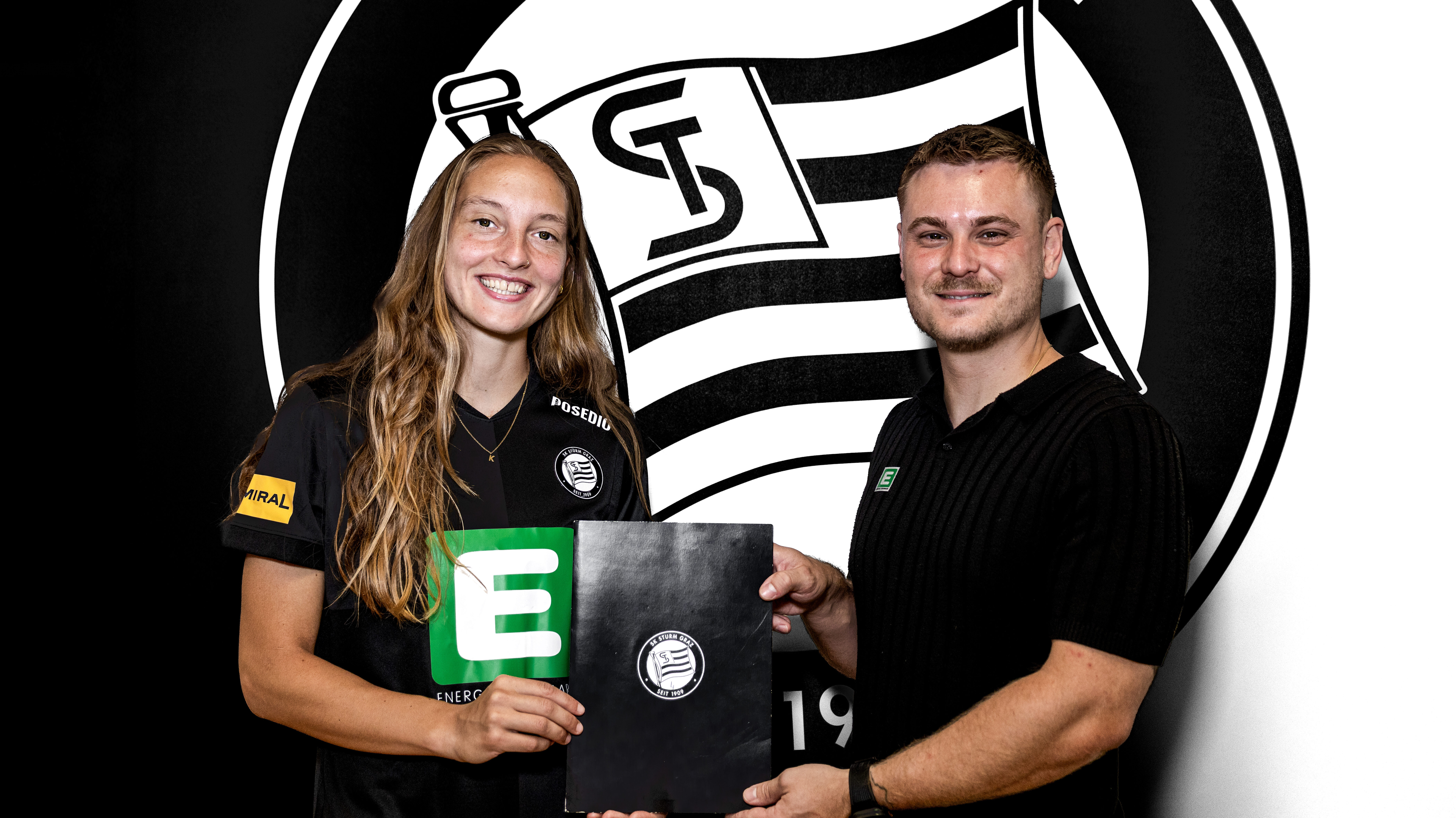 Eine Frau und ein Mann in einem schwarzen Shirt stehen vor einem schwarz-weißen Logo und halten eine schwarze Mappe.