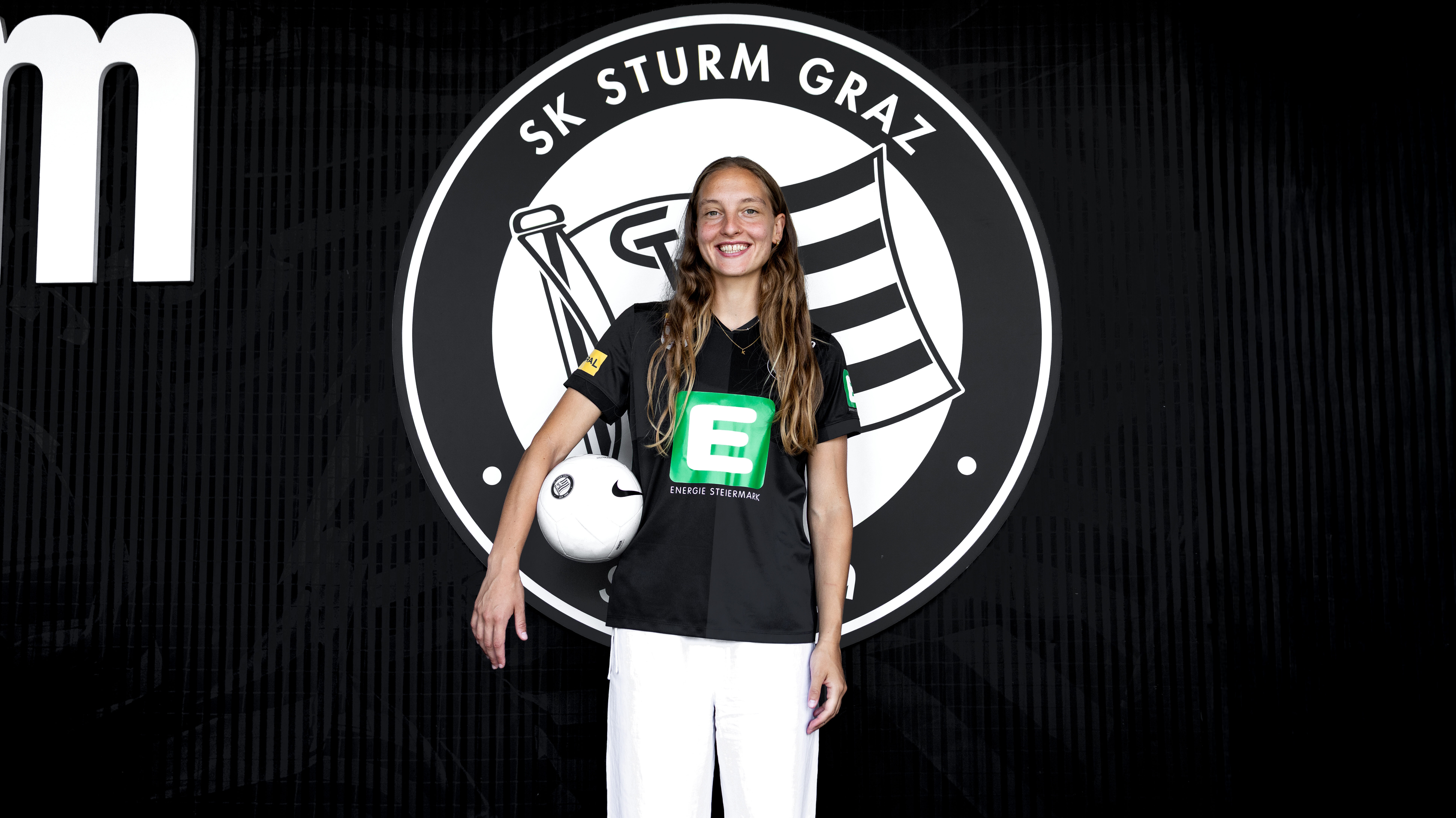 Eine Frau in einem schwarzen Shirt und einer weißen Hose steht vor einem schwarz-weißen Logo und hält einen weißen Ball in der Hand.