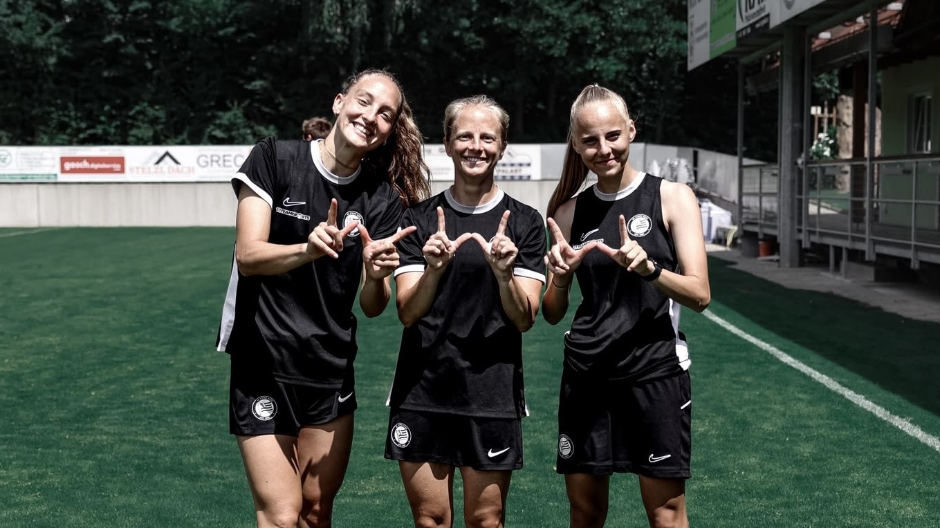 Drei Frauen in schwarzen Shirts lächeln und zeigen ein W.