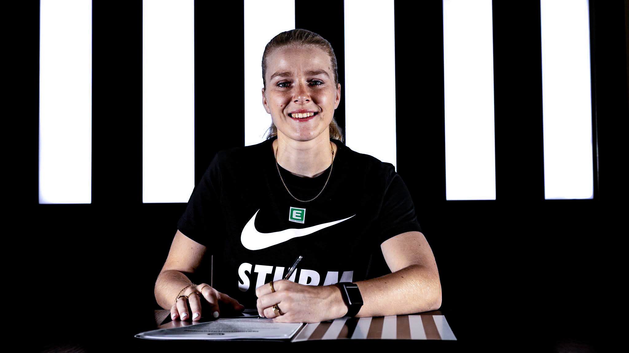 Eine Spielerin sitzt in einem schwarzen Shirt vor einer schwarz-weißen Wand und unterschreibt einen weißen Zettel.