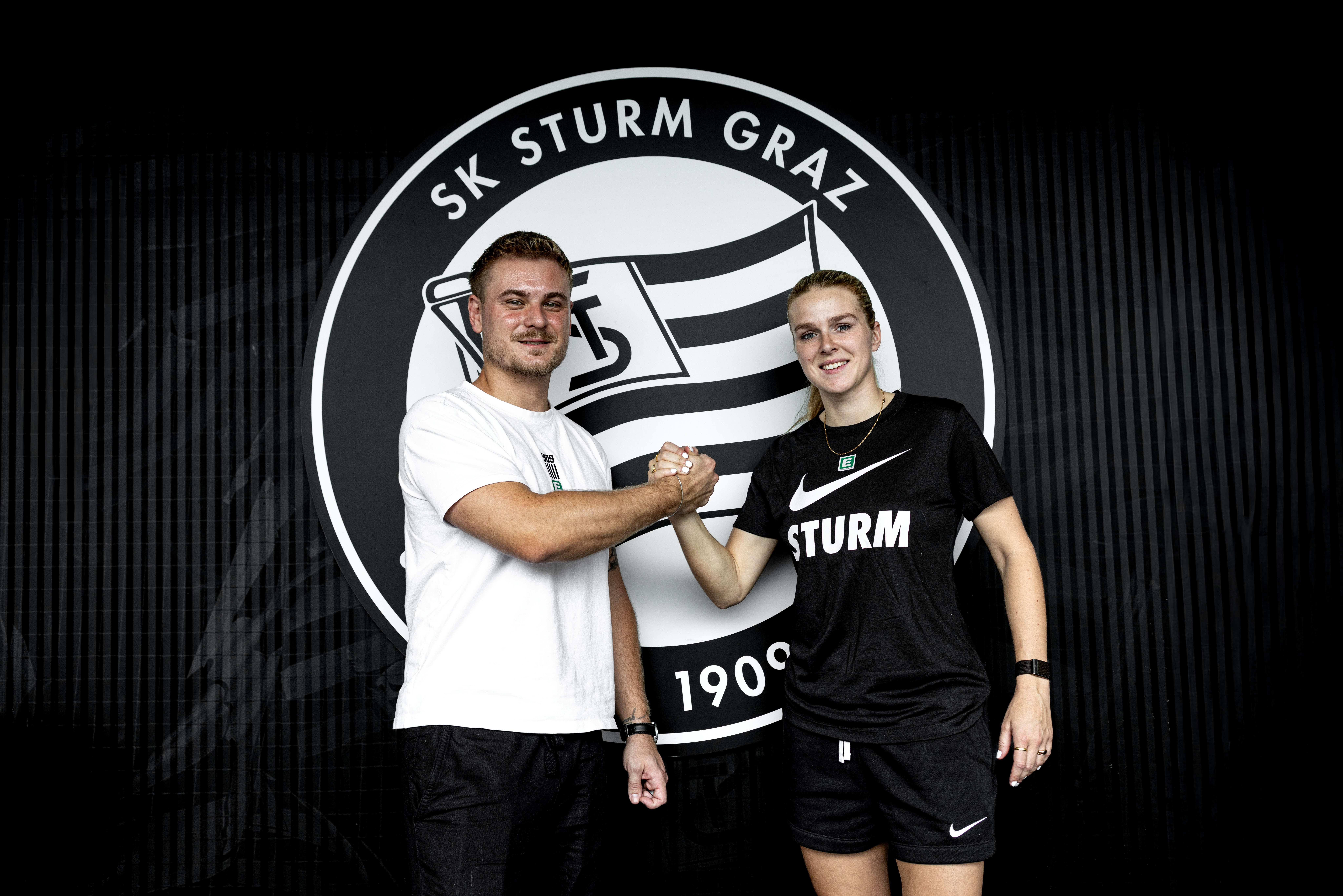 Ein Mann in einem weißen Shirt und eine Frau in einem schwarzen klatschen vor einem schwarz-weißen Logo ein