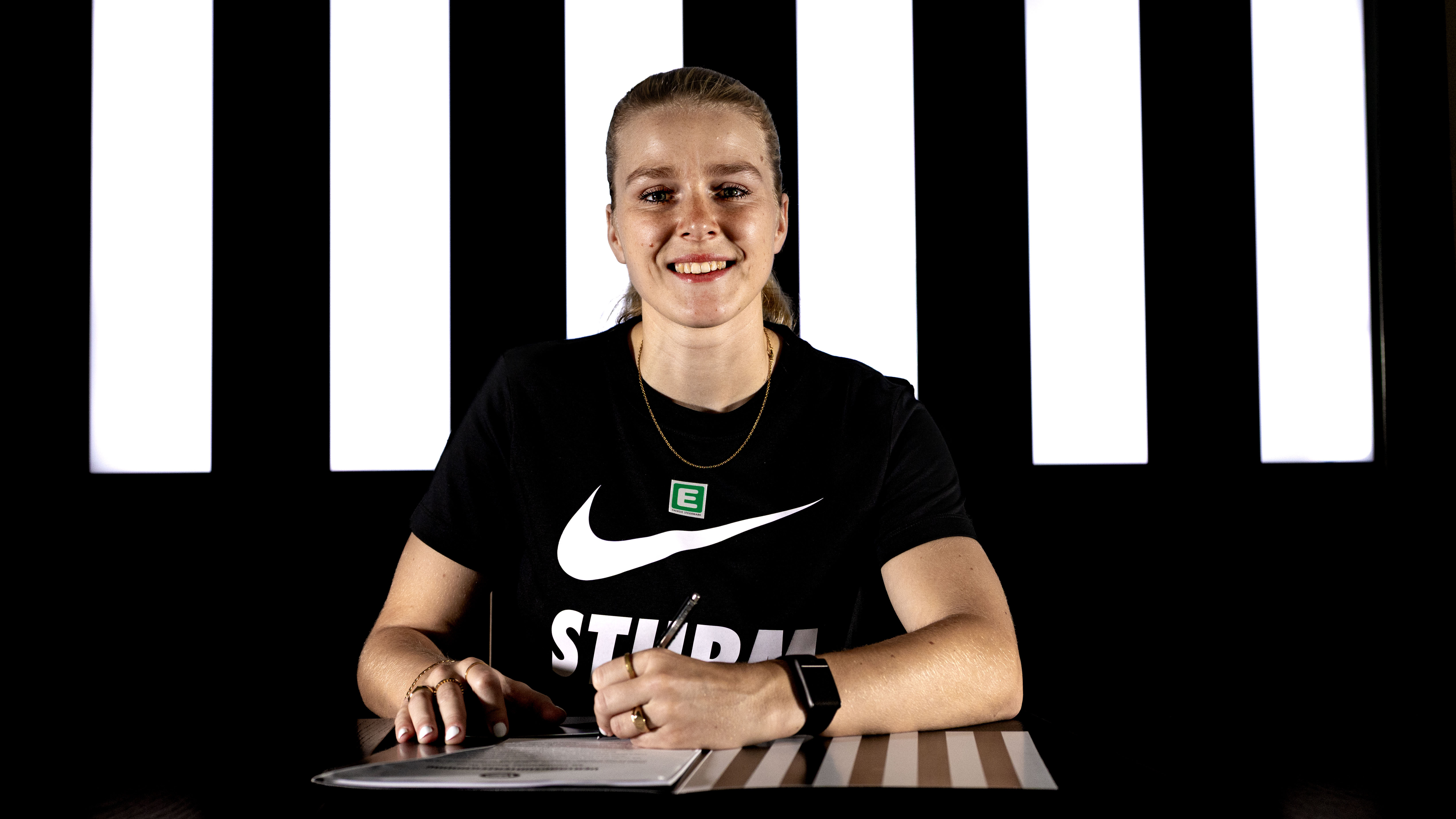 Eine Spielerin sitzt in einem schwarzen Shirt vor einer schwarz-weißen Wand und unterschreibt einen weißen Zettel.