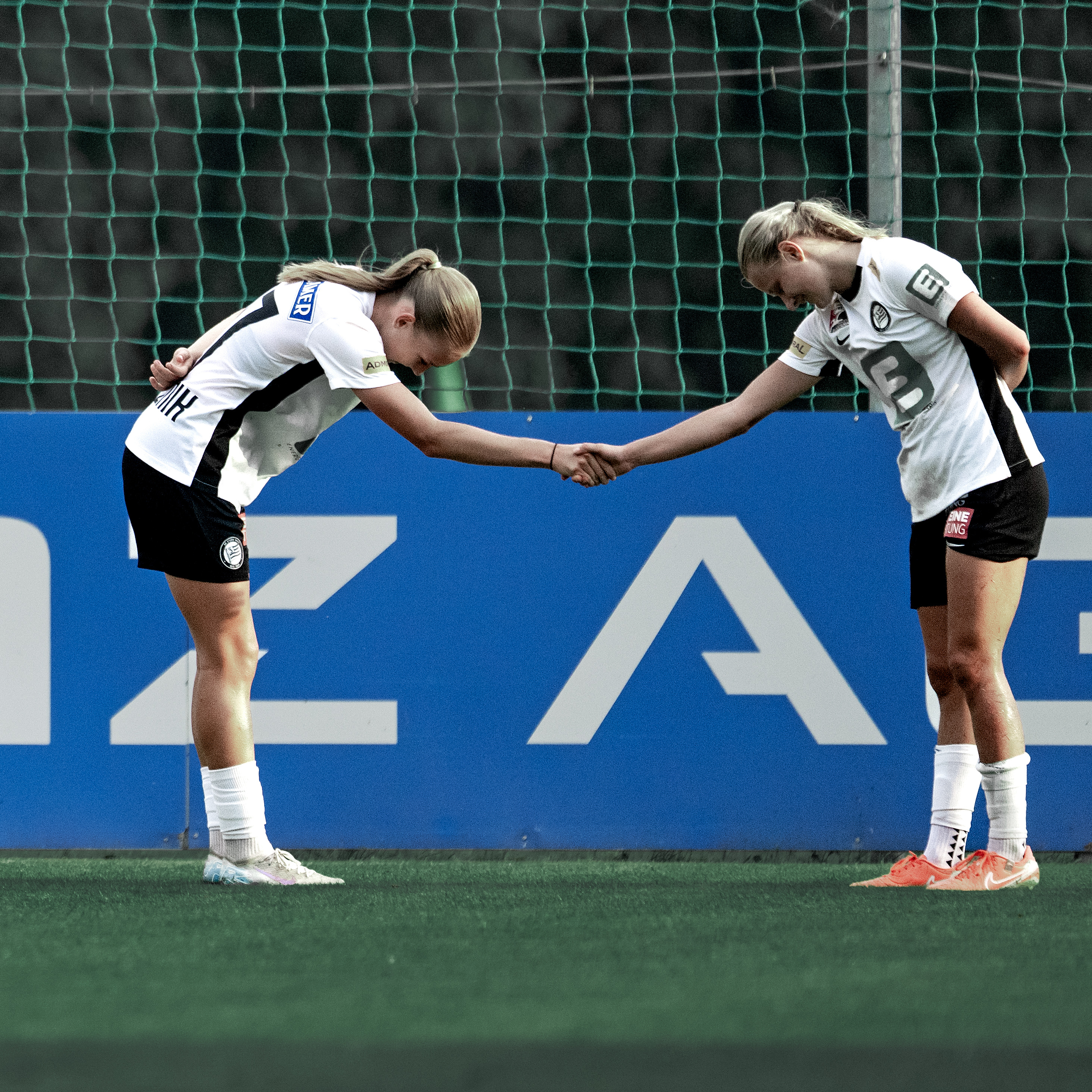 Zwei Frauen in weißen Shirts die sich die Hand geben und nach vorne beugen.