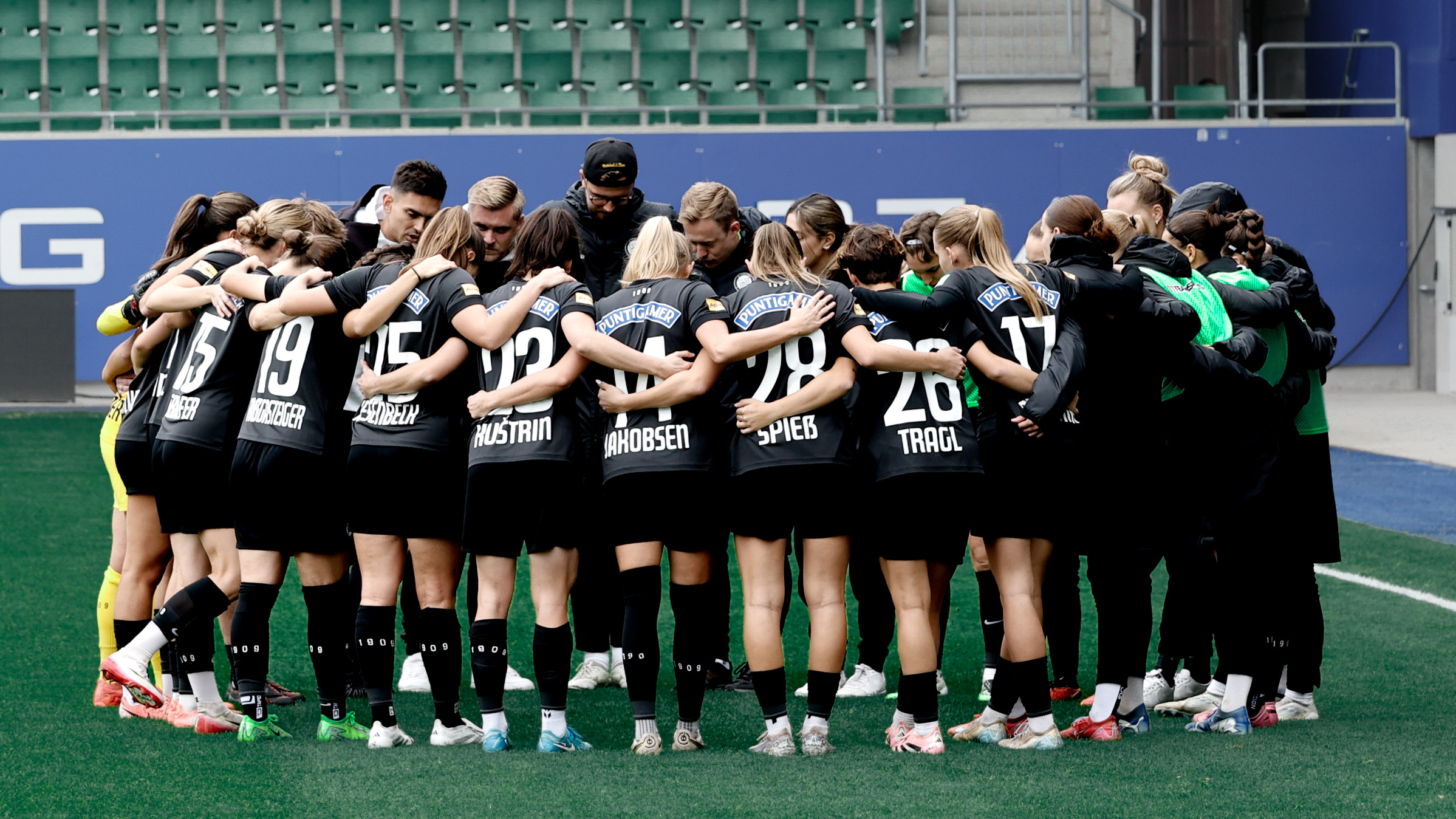 Eine Gruppe voller Frauen mIt schwarzem Trikot stehen im Kreis zusammen und Umarmen sich.