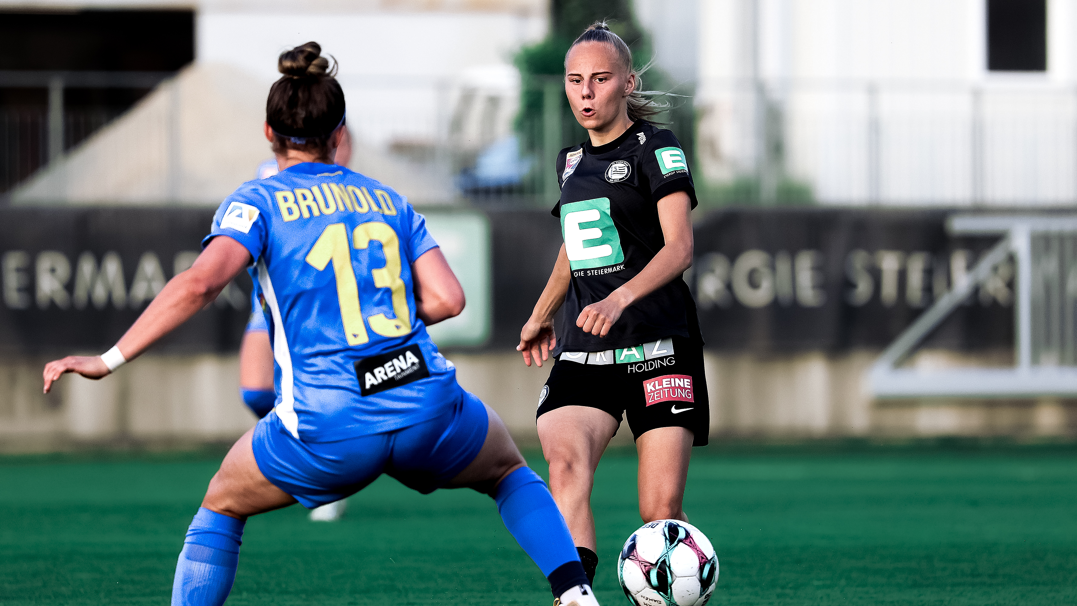 Eine Spielerin in schwarz tritt gegen einen schwarz-weißen Ball und eine Spielerin in blau versucht den Ball abzufangen.
