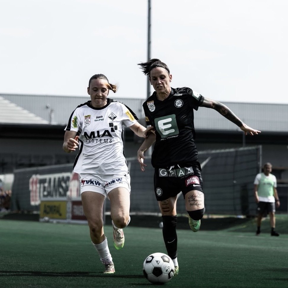 Eine Frau in weißem Trikot und eine Frau in schwarzem Trikot laufen den Ball hinterher.