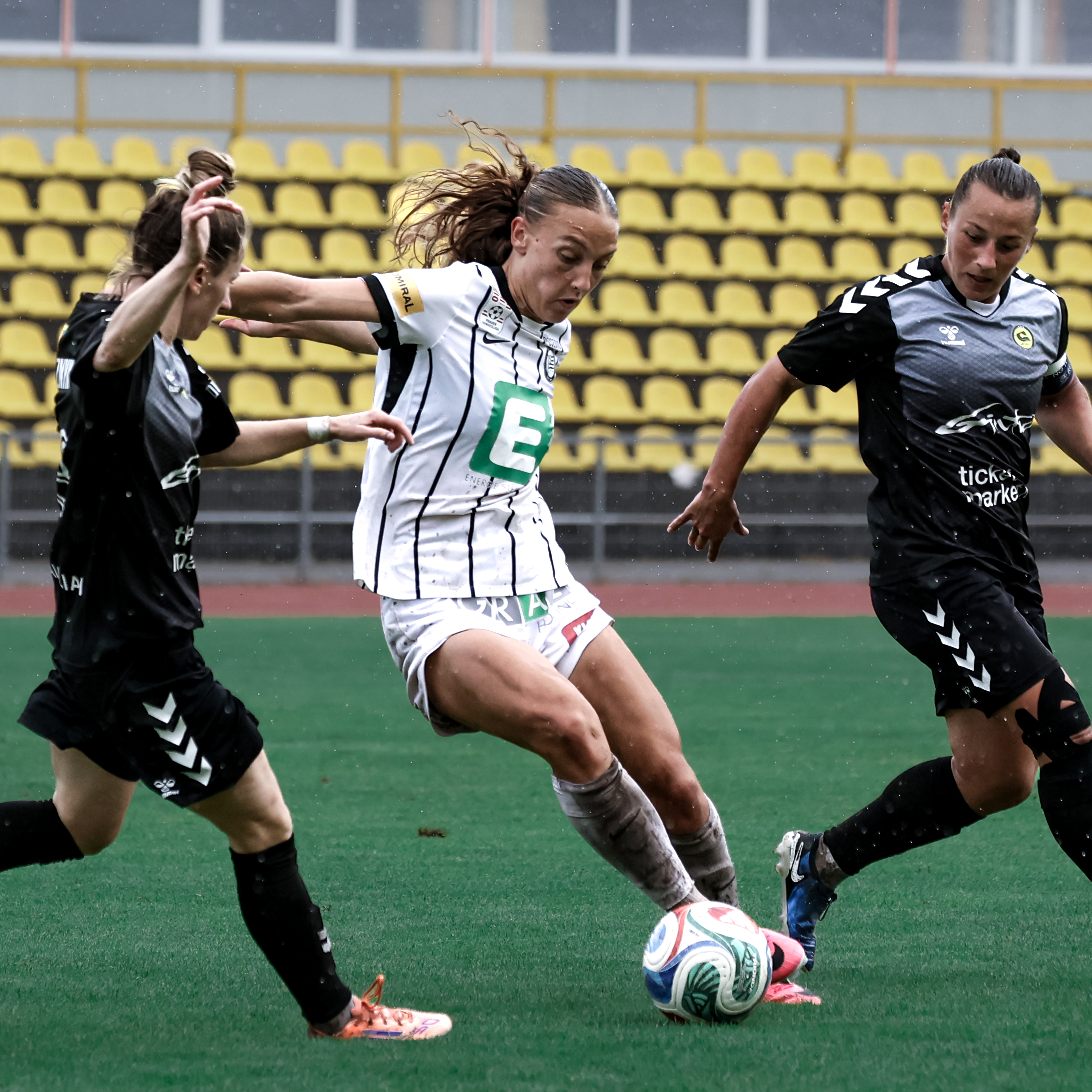 Eine Frau in weißem Trikot hat den Ball am Fuß und zwei mit schwarzen Trikots laufen neben ihr.