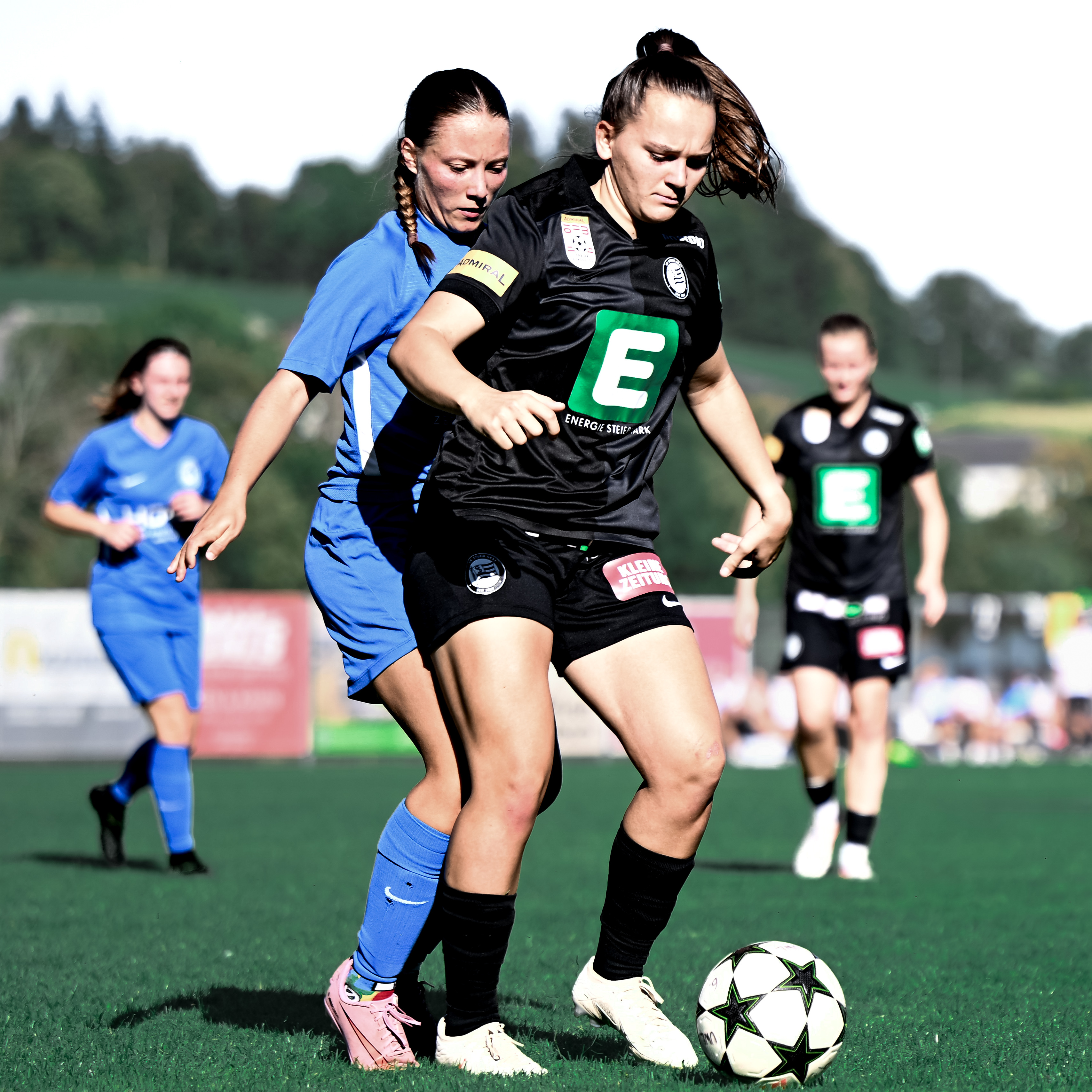 Eine Frau in schwarzem Trikot hat den Ball am Fuß eine Frau in blauem Trikot steht hinter ihr.