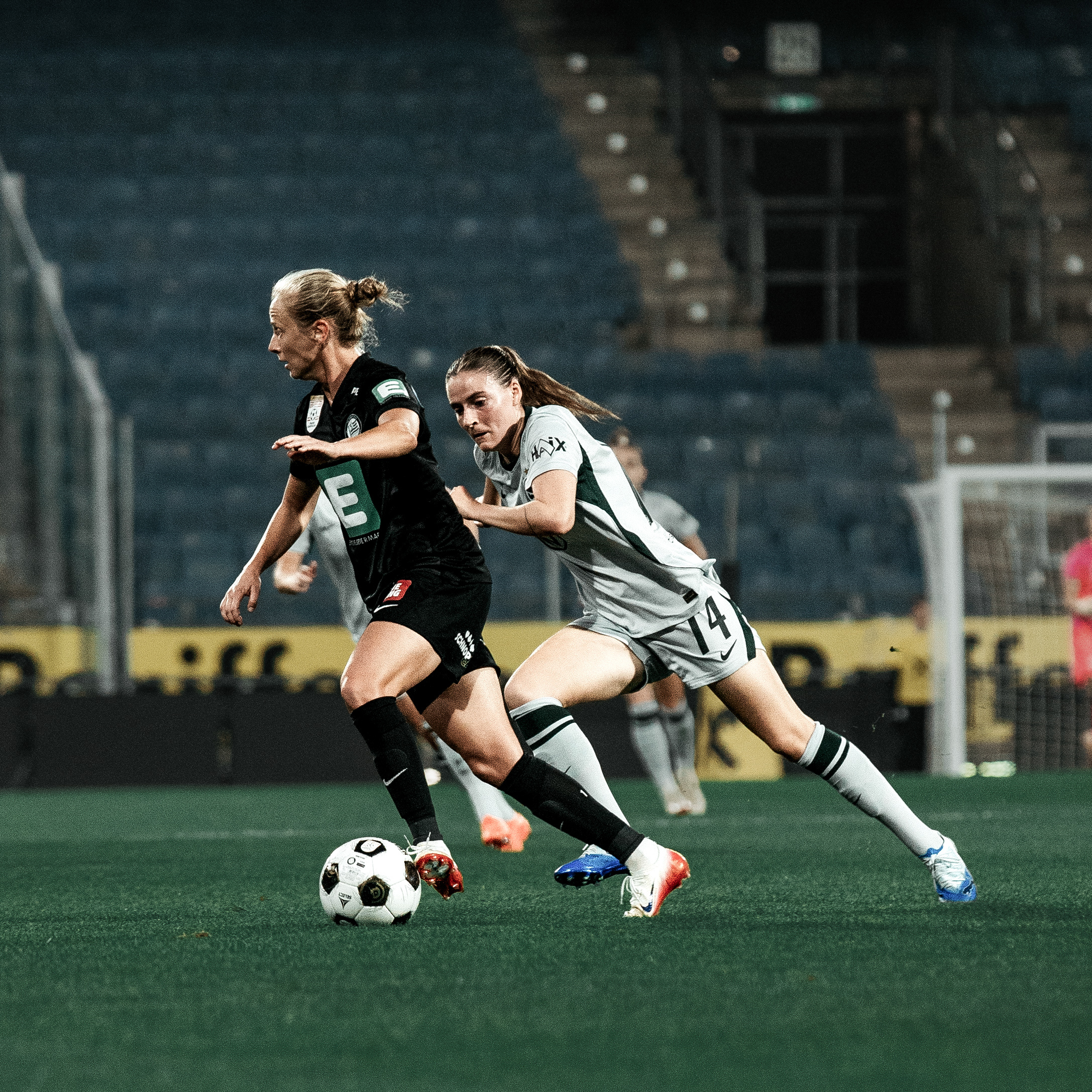 Eine Frau in schwarzem Trikot hat den Ball am Fuß, eine Frau in grauem Trikot läuft ihr hinterher.