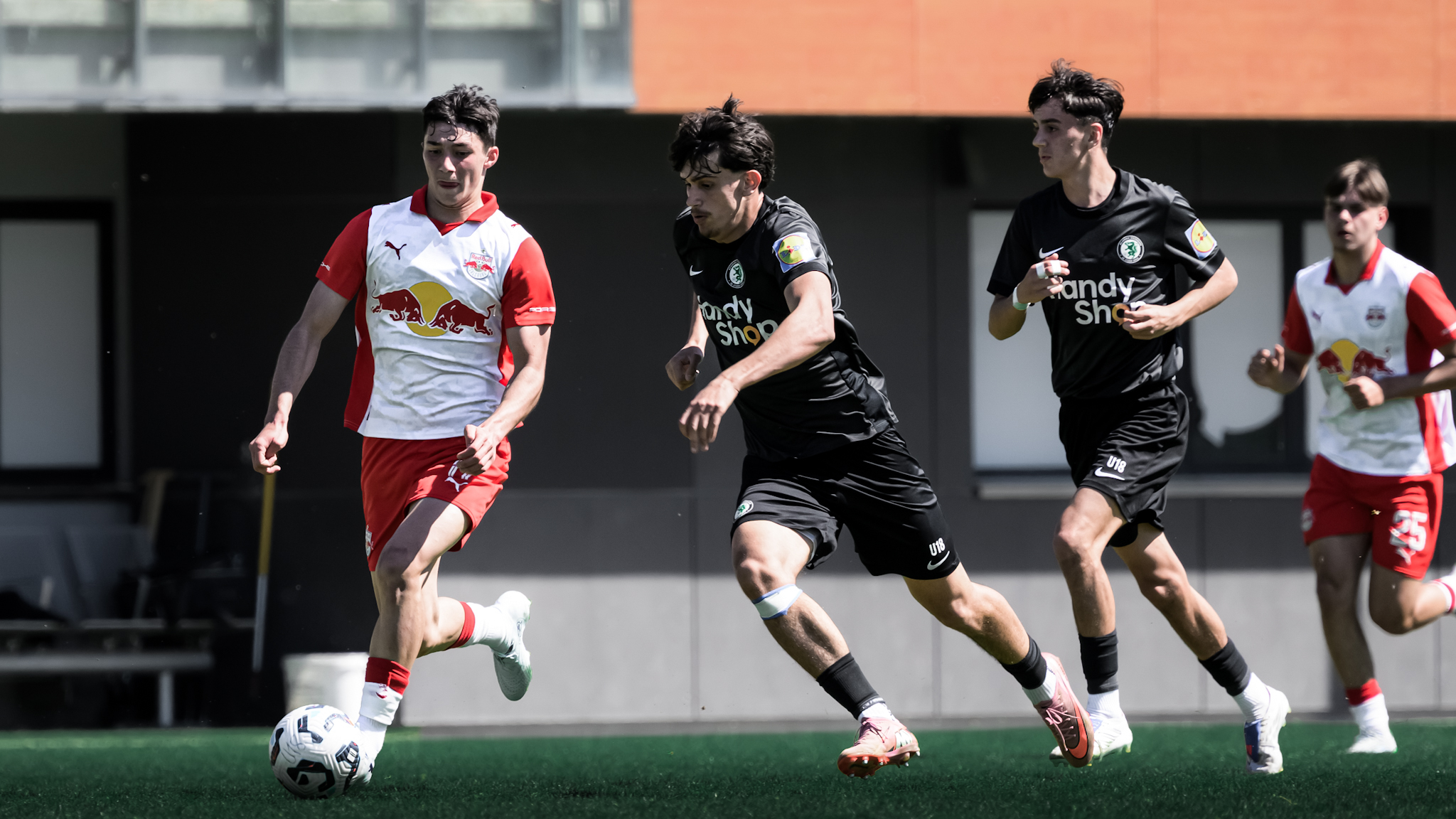 Ein Spieler in schwarz und eine Spieler in rot-weiß kämpfen um einen weißen Ball. Im Hintergrund sind noch ein weiterer Spieler in schwarz und einer in rot-weiß zu sehen.