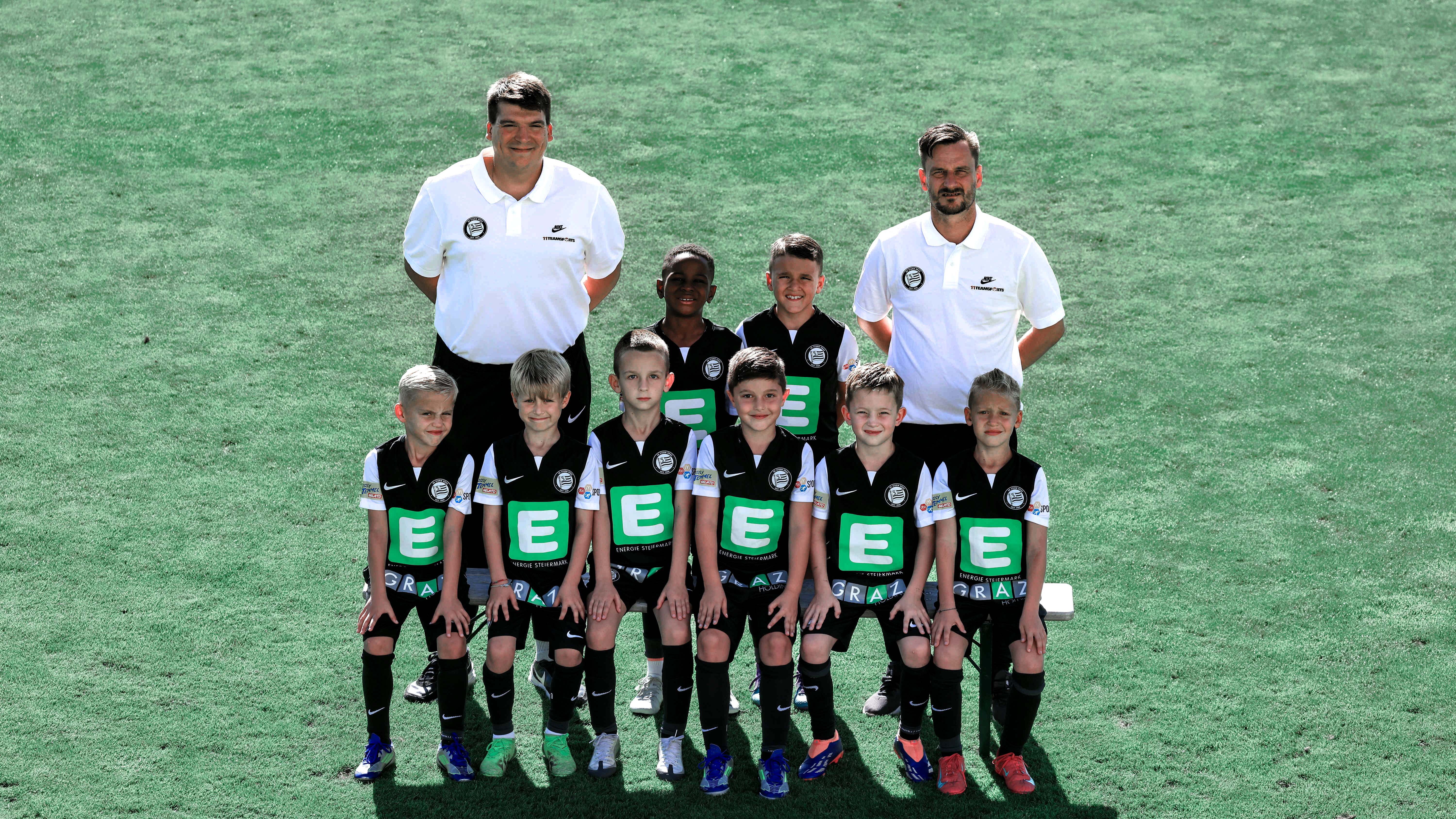 Viele Kinder in schwarzen Trikots stehen in mehreren Reihen und einige erwachsene Trainer haben weiße Shirts an