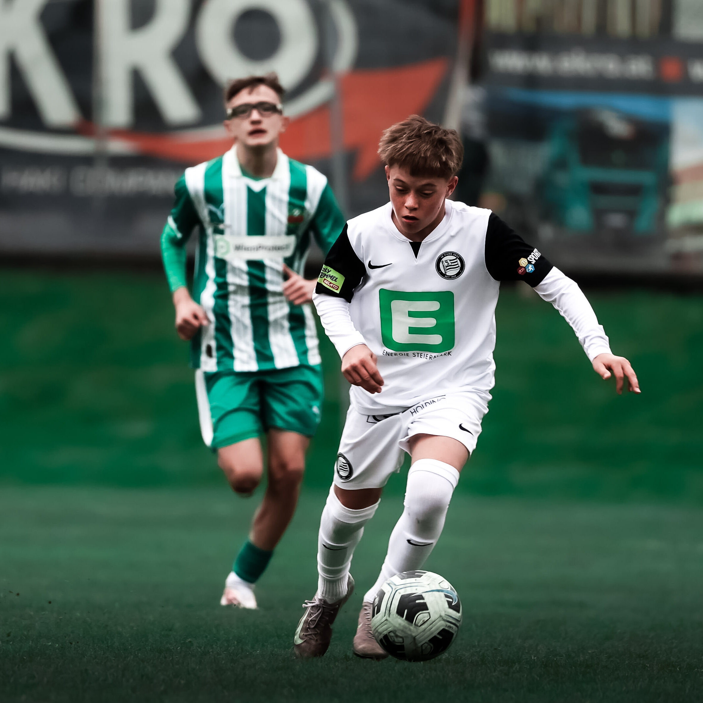 Ein junger Spieler in weißem Trikot hat einen weißen Ball am Fuß und wird von einem jungen Spieler in grün-weiß verfolgt