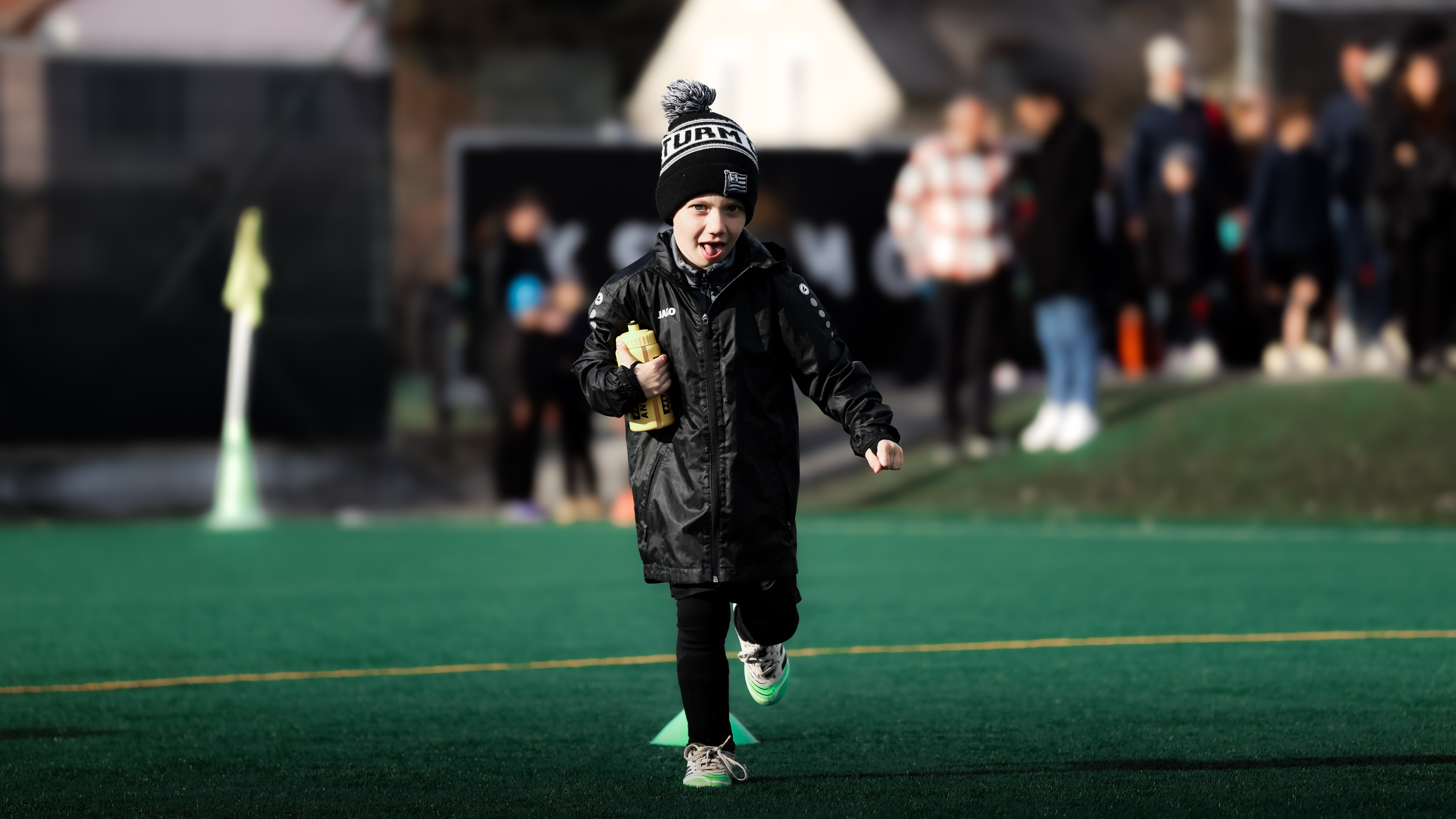 Ein Kind mit einer Trinkflasche und in schwarz gekleidet läuft über Kunstrasen