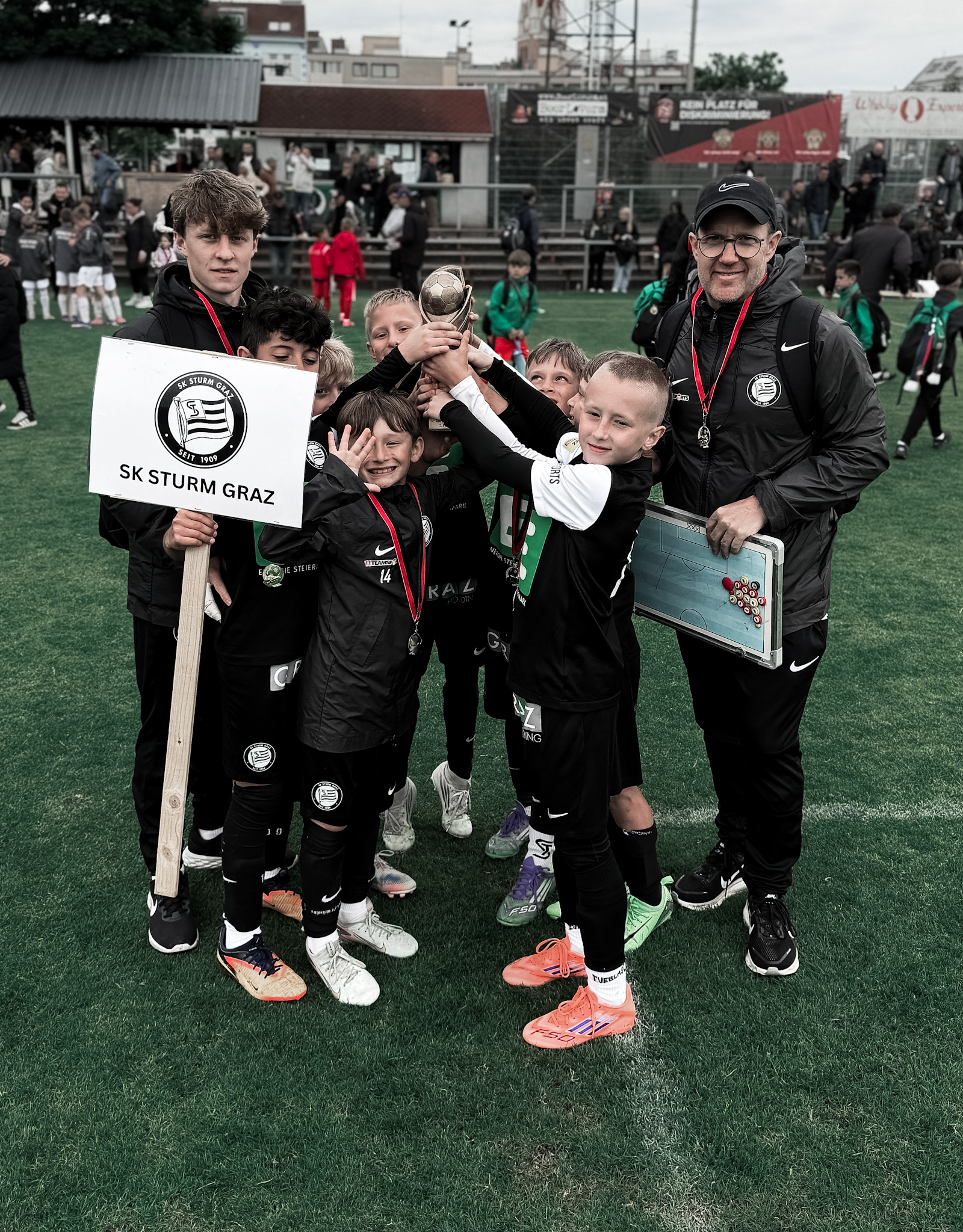 Viele junge Spieler halten einen Pokal hoch, zwei Trainer stehen seitlich dahinter