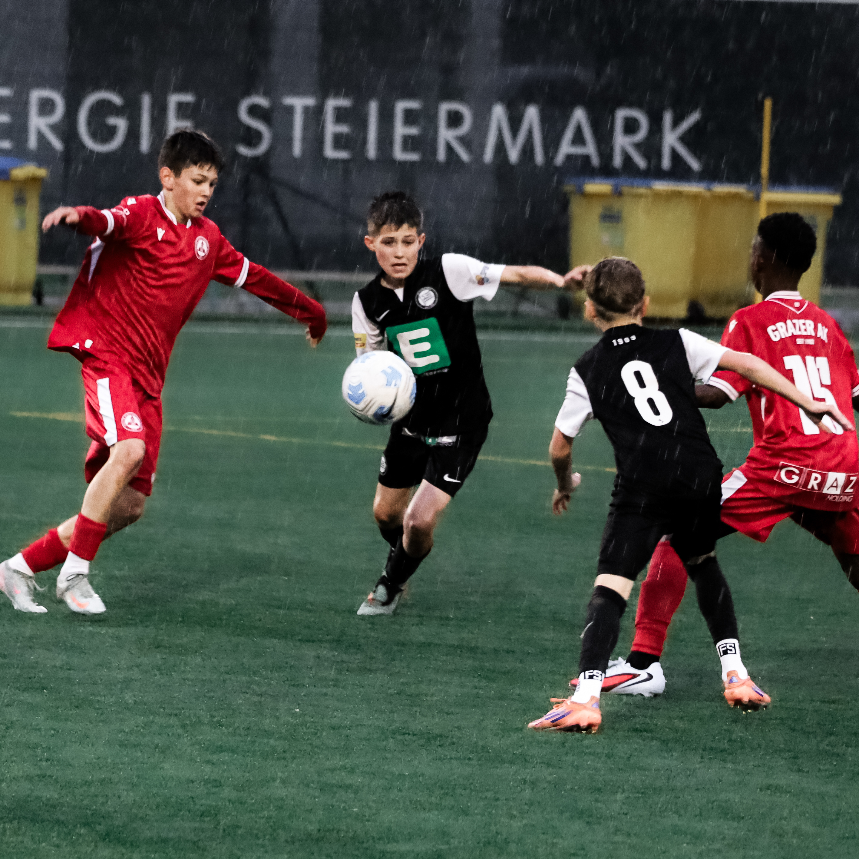 Zwei Spieler in schwarzen Trikots kaempfen mit zwei Spielern in roten Trikots um einen weißen Ball