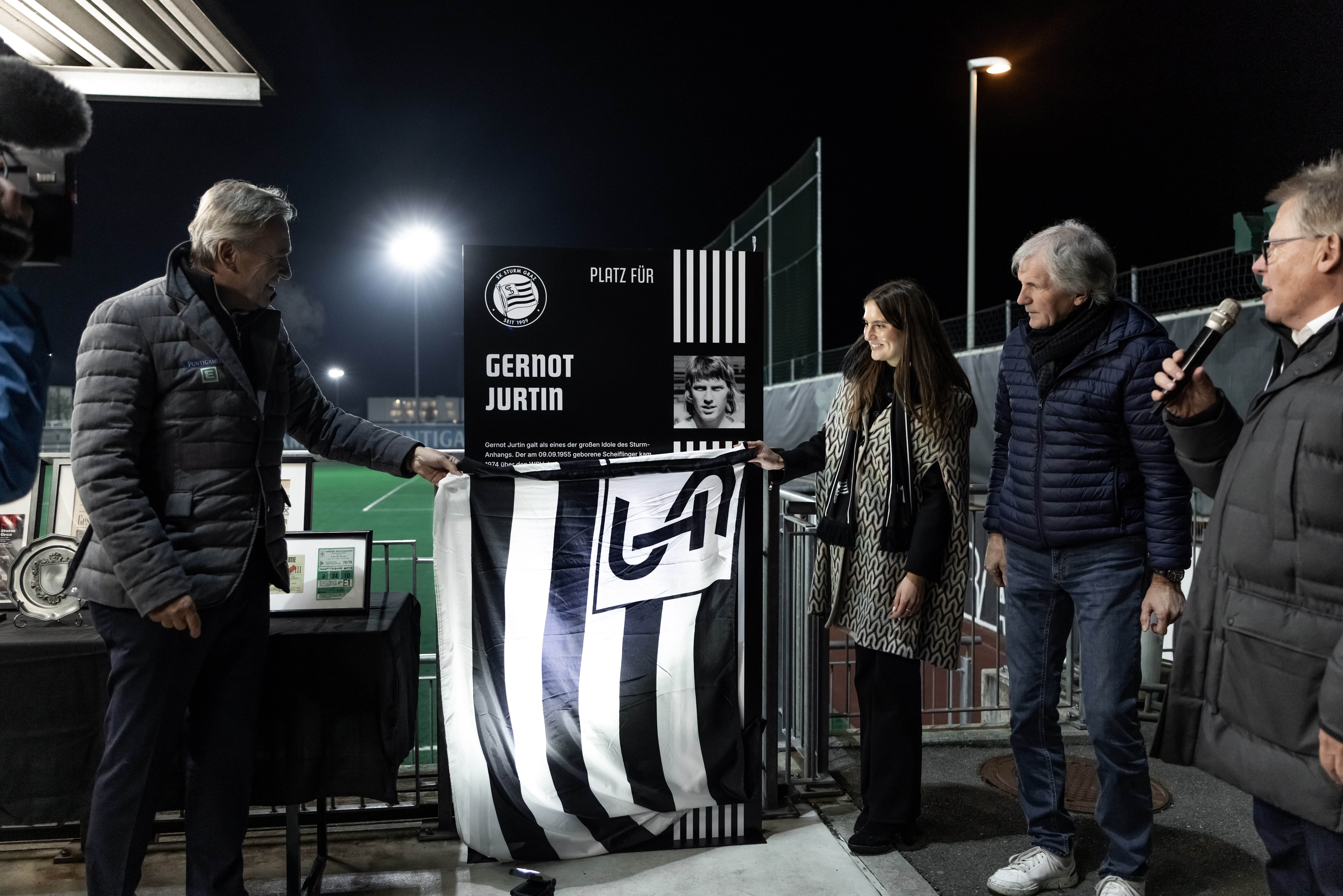 Ein Mann und eine Frau enthüllen eine Stele, die unter einer Fahne hervorkommt