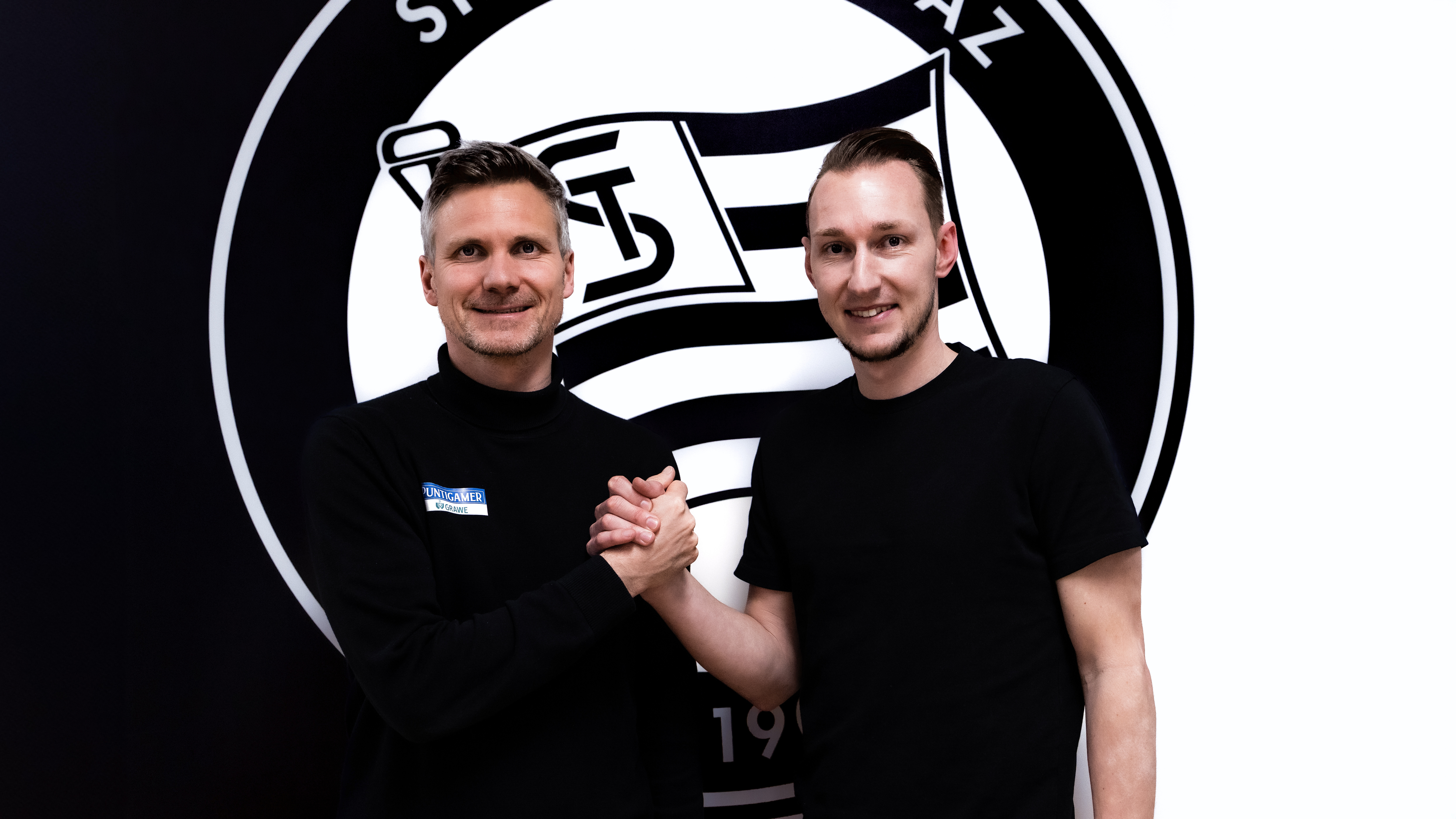 Zwei Männer geben sich vor einem Logo die Hände