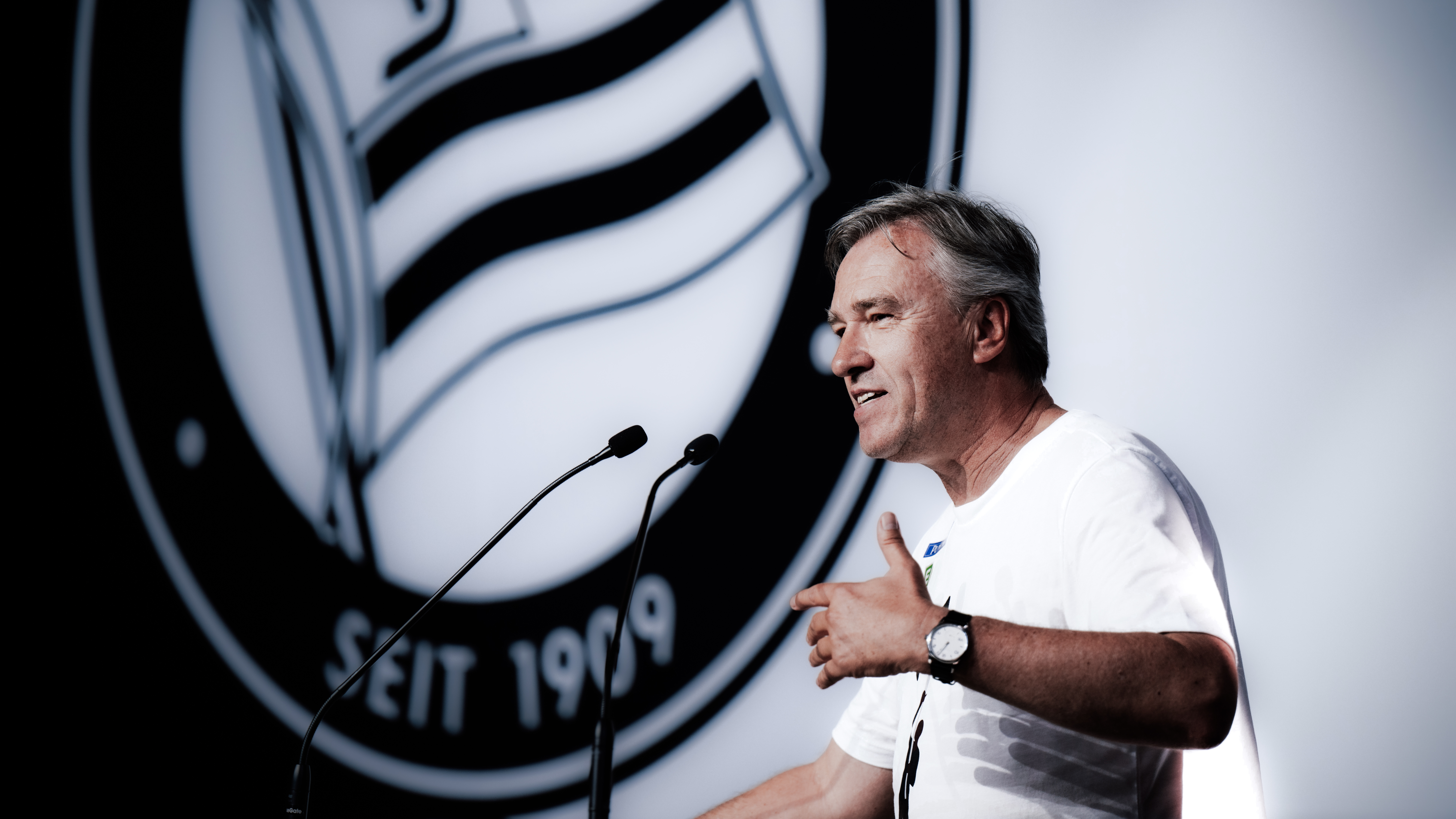 Ein Mann in einem weißen Shirt steht vor einem Logo und spricht in Mikrofone