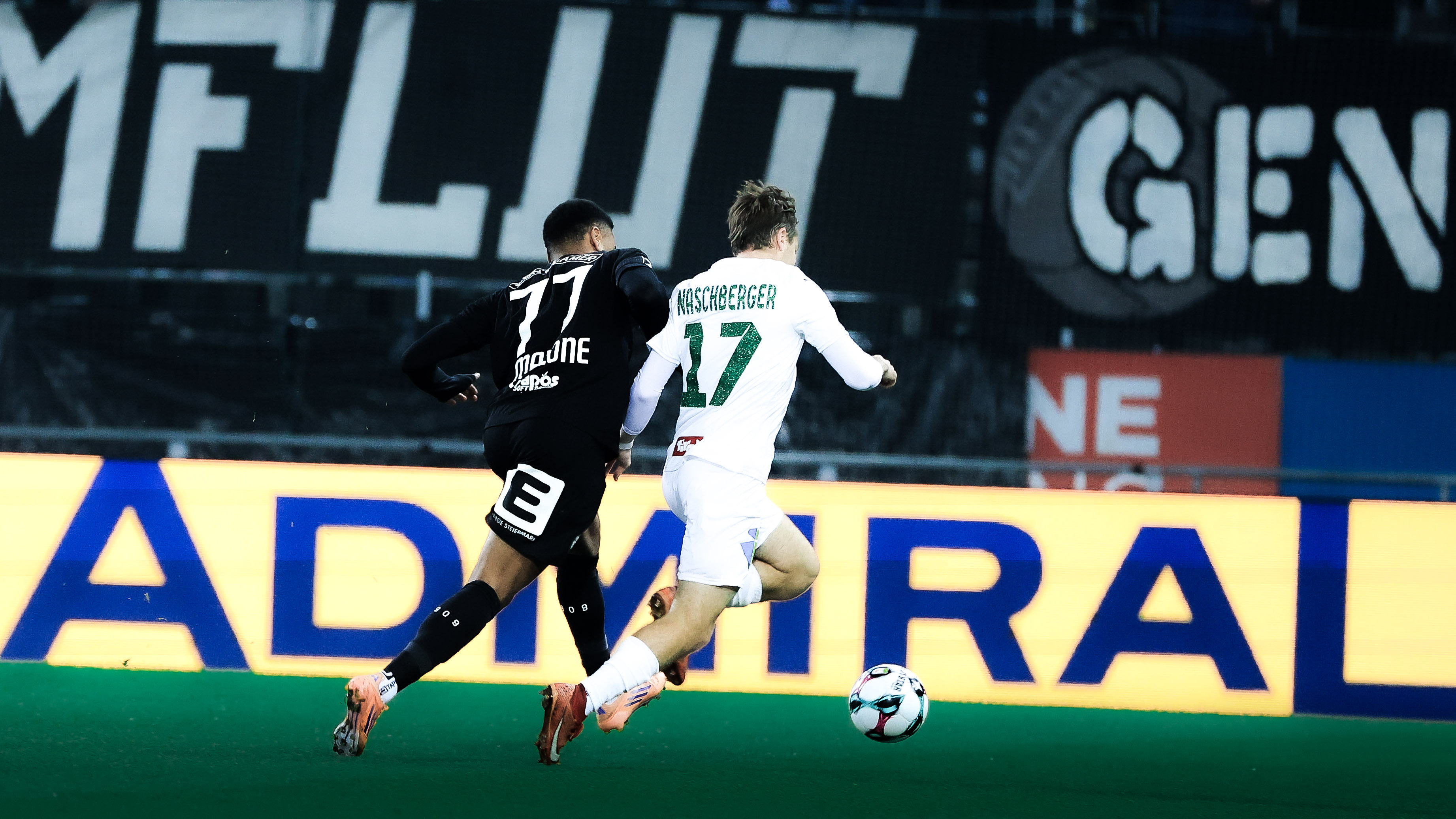 Zwei Spieler – einer in schwarz und einer in weiß – kämpfen vor einer gelben LED Bande um einen weißen Ball.