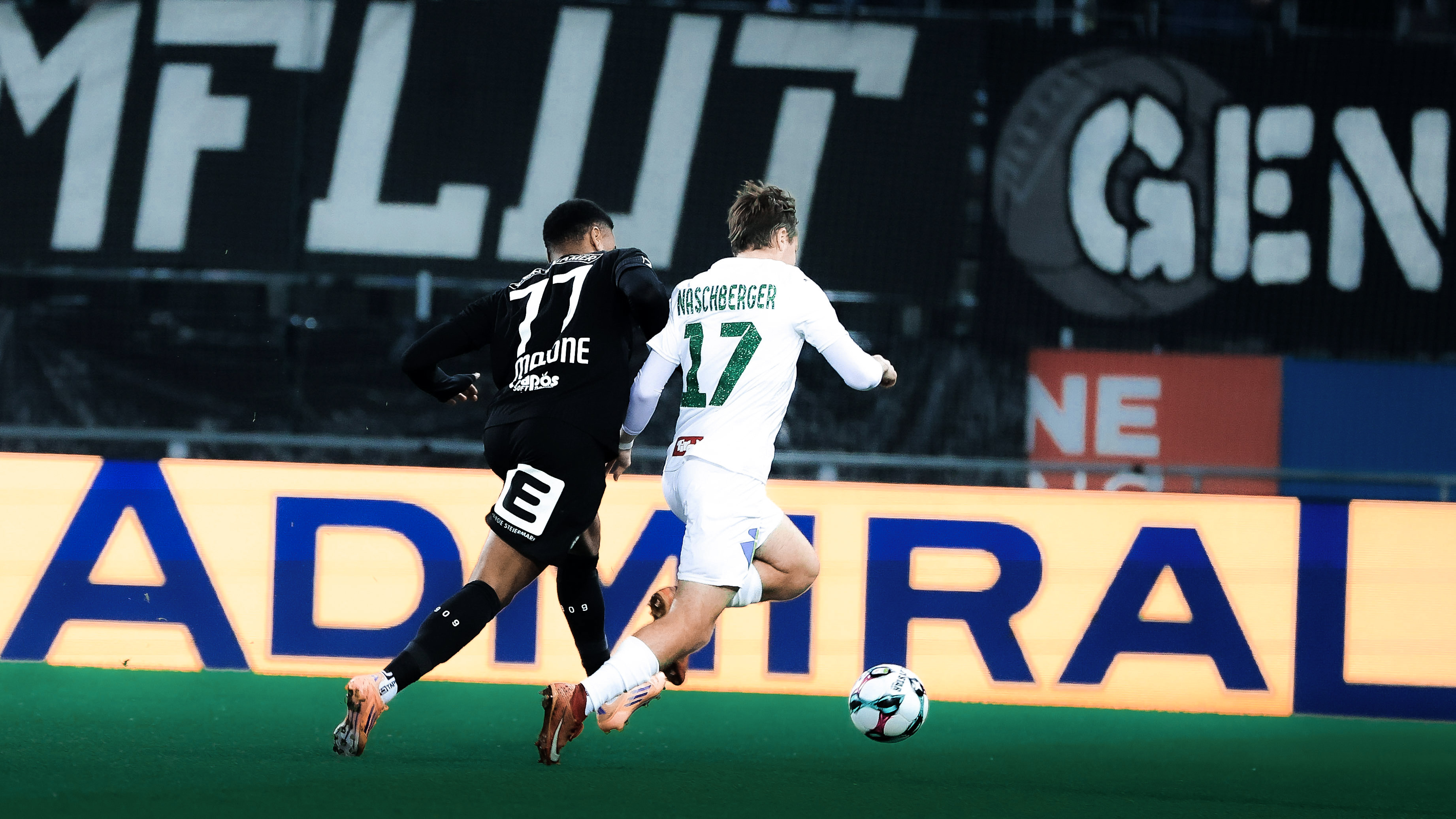 Zwei Spieler – einer in schwarz und einer in weiß – kämpfen vor einer gelben LED Bande um einen weißen Ball.