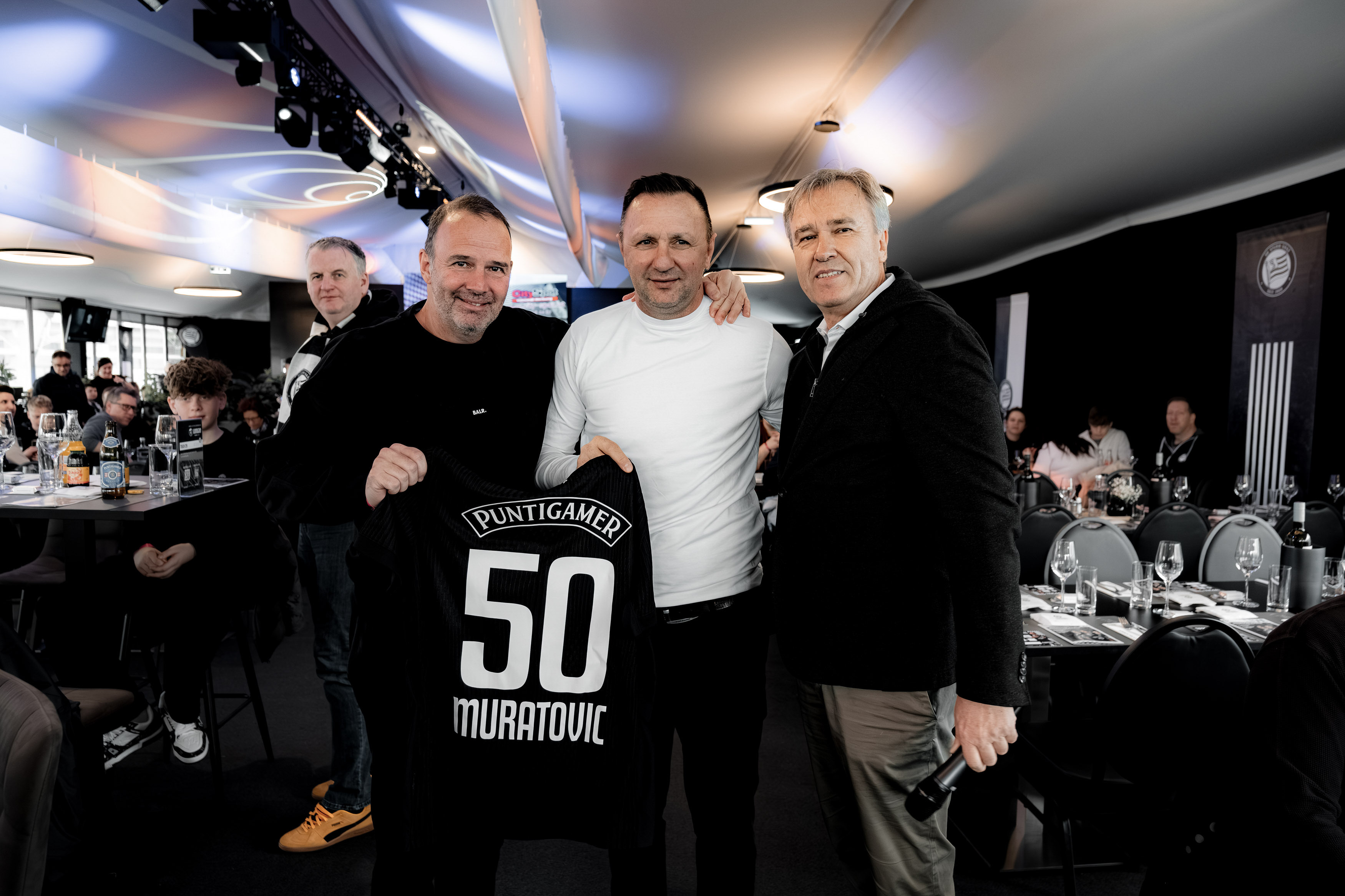 Drei Männer halten ein schwarzes Trikot mit der 50