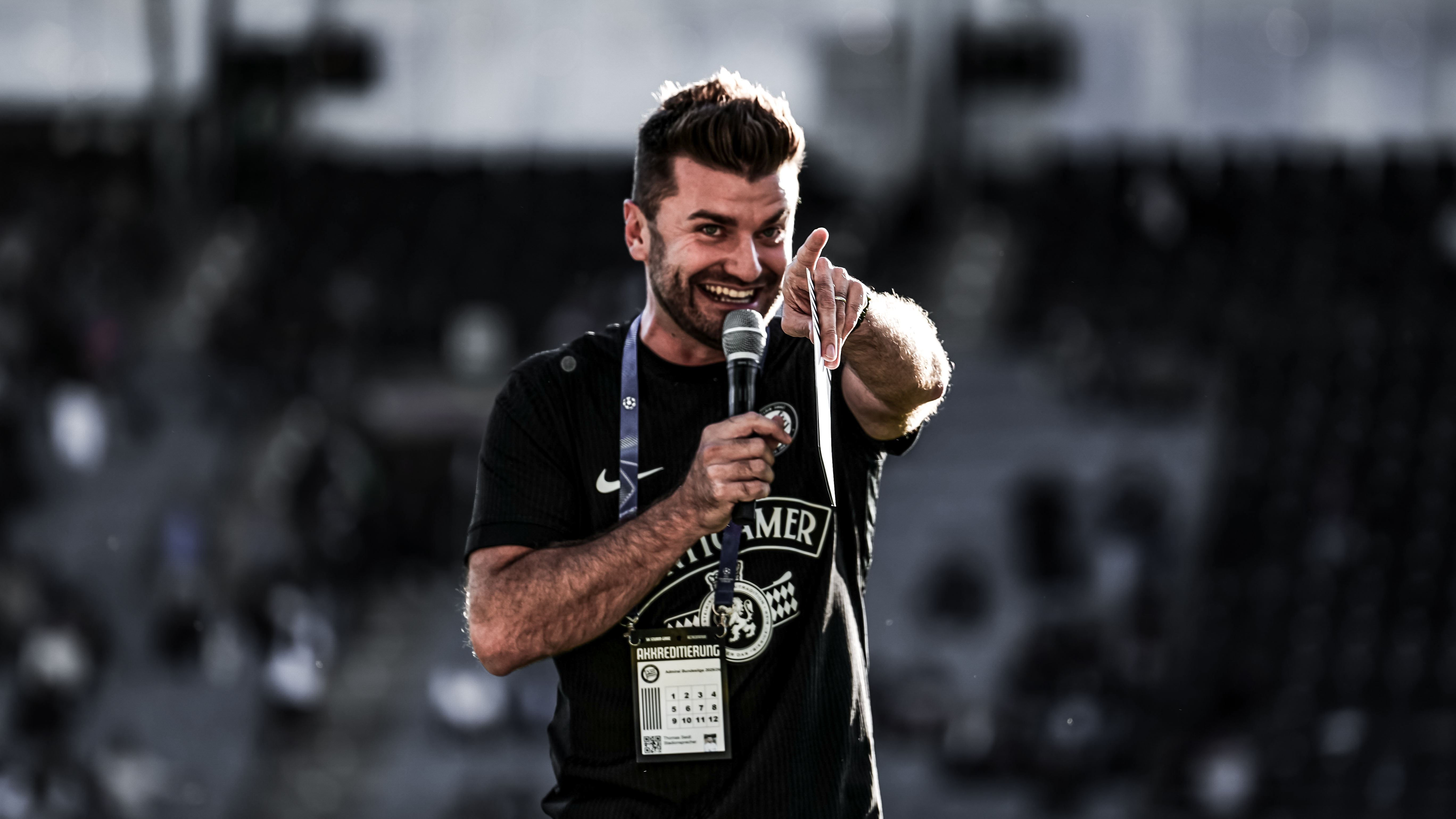 Ein Mann in einem schwarzen Trikot zeigt in eine Richtung mit einem Mikro in der Hand