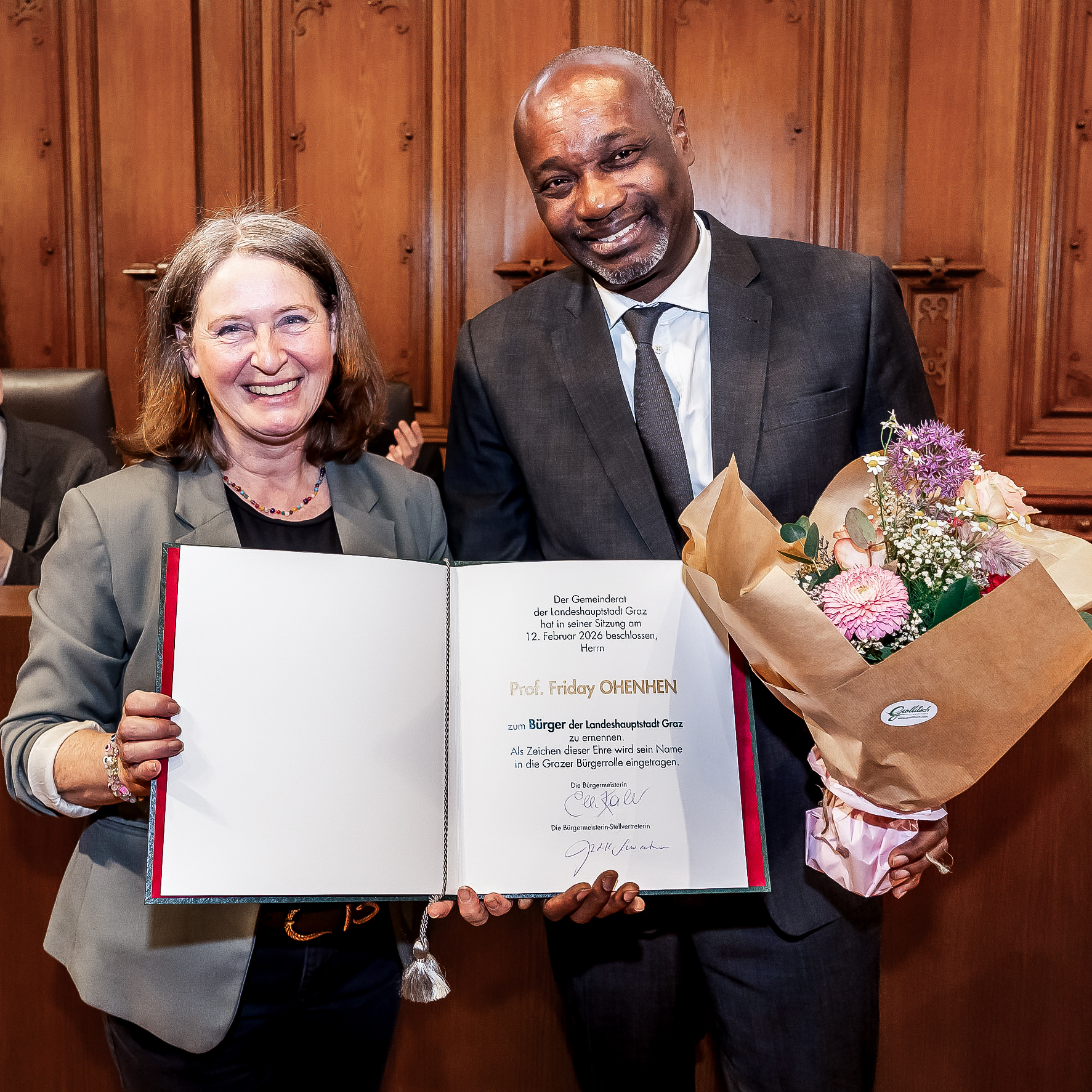 Ein Mann im Anzug und eine Frau im Blazer stehen nebeneinander, halten ein Buch und einen Blumenstrauß in der Hand.