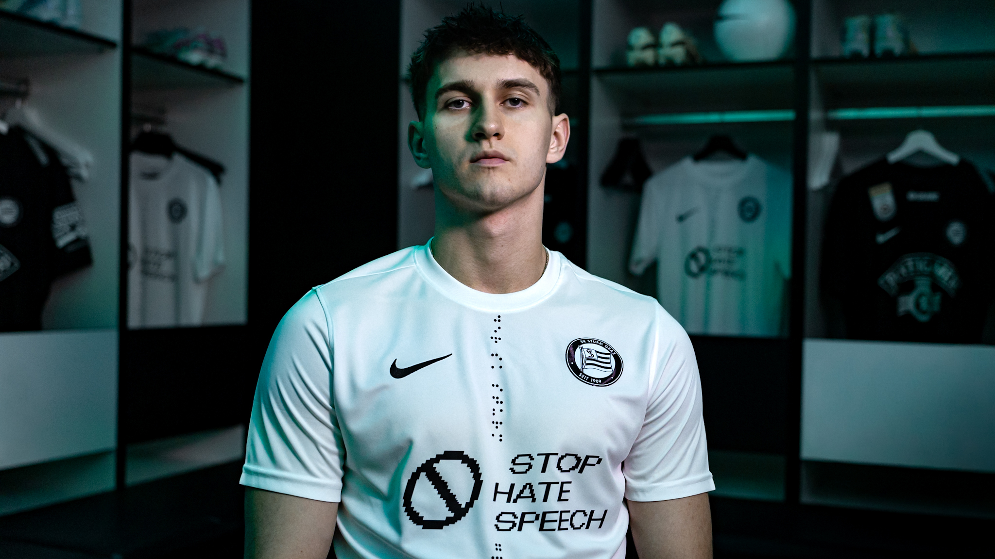 Eine männliche Person sitzt in einer Kabine und trägt ein weißes Trikot mit der Aufschrift "Stop Hate Speech"