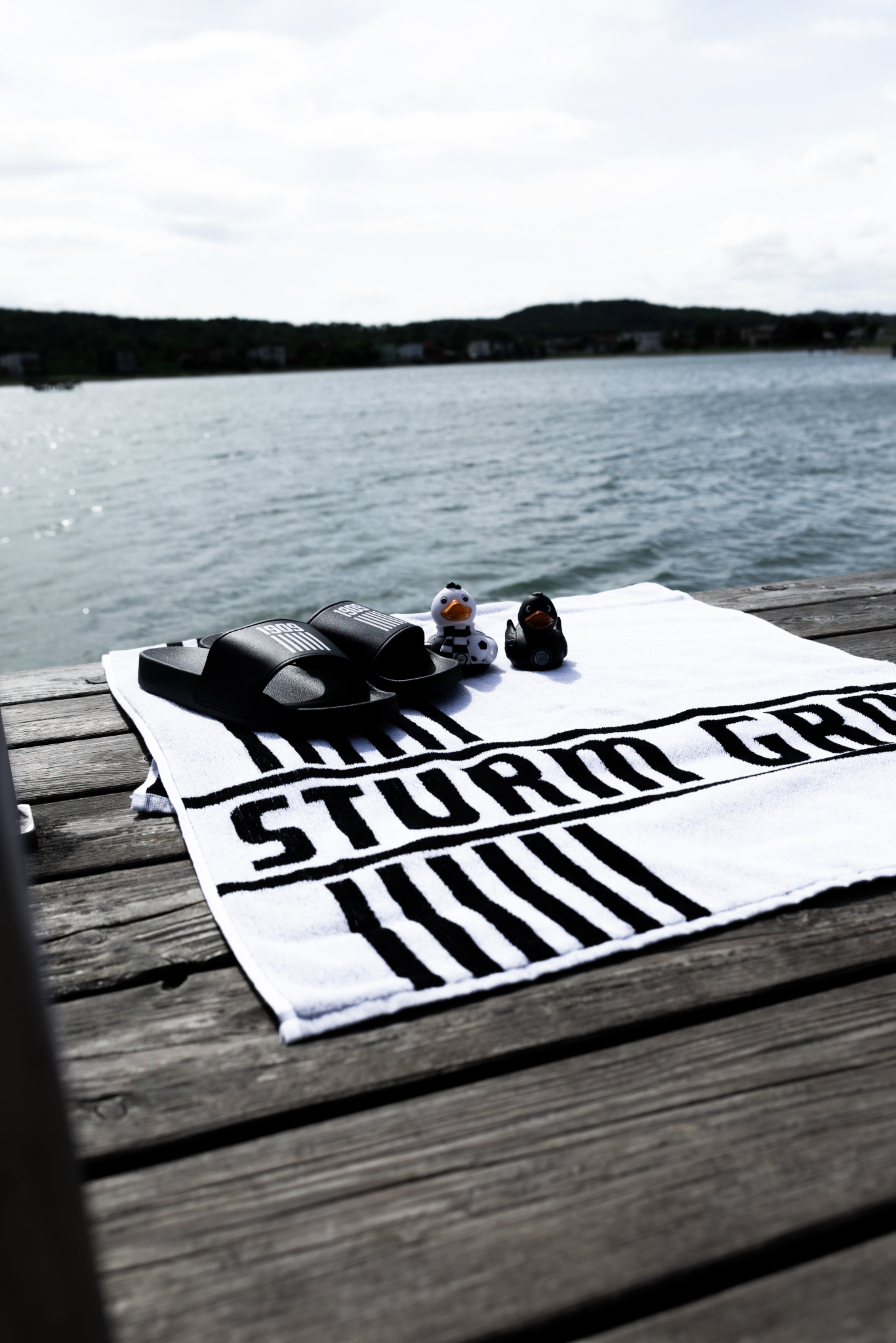 Ein Handtuch mit Badeschlappen und Badeente auf einem Steg vor einem See.