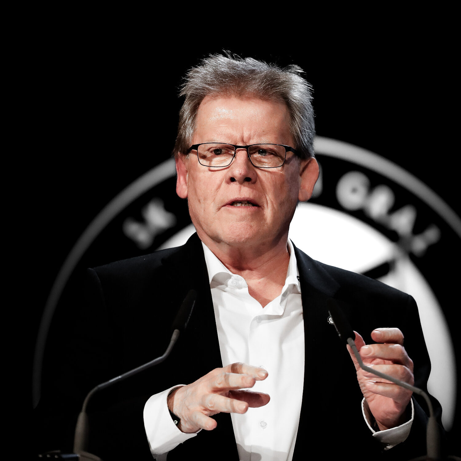 Ein älterer Mann mit Brille steht vor einem Pult und spricht.