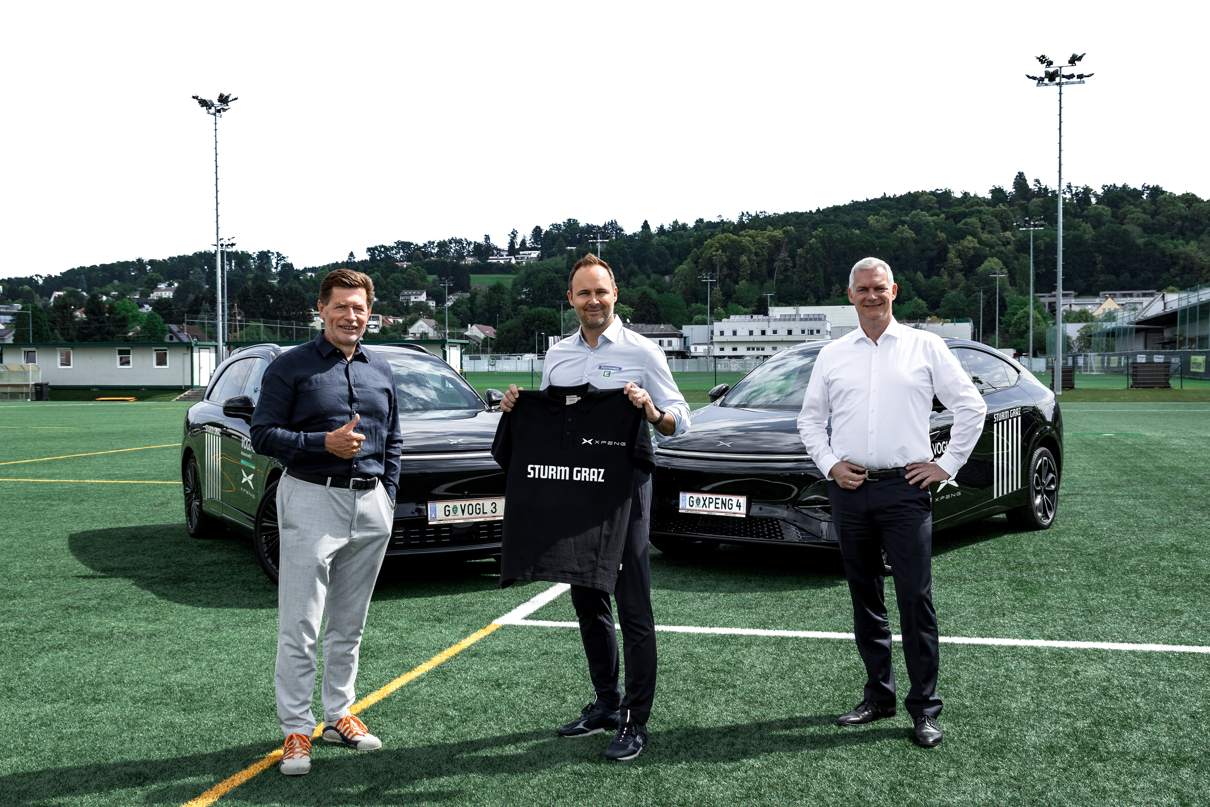 Drei Männer halten ein T-Shirt in die Kamera und stehen vor zwei schwarzen Autos.