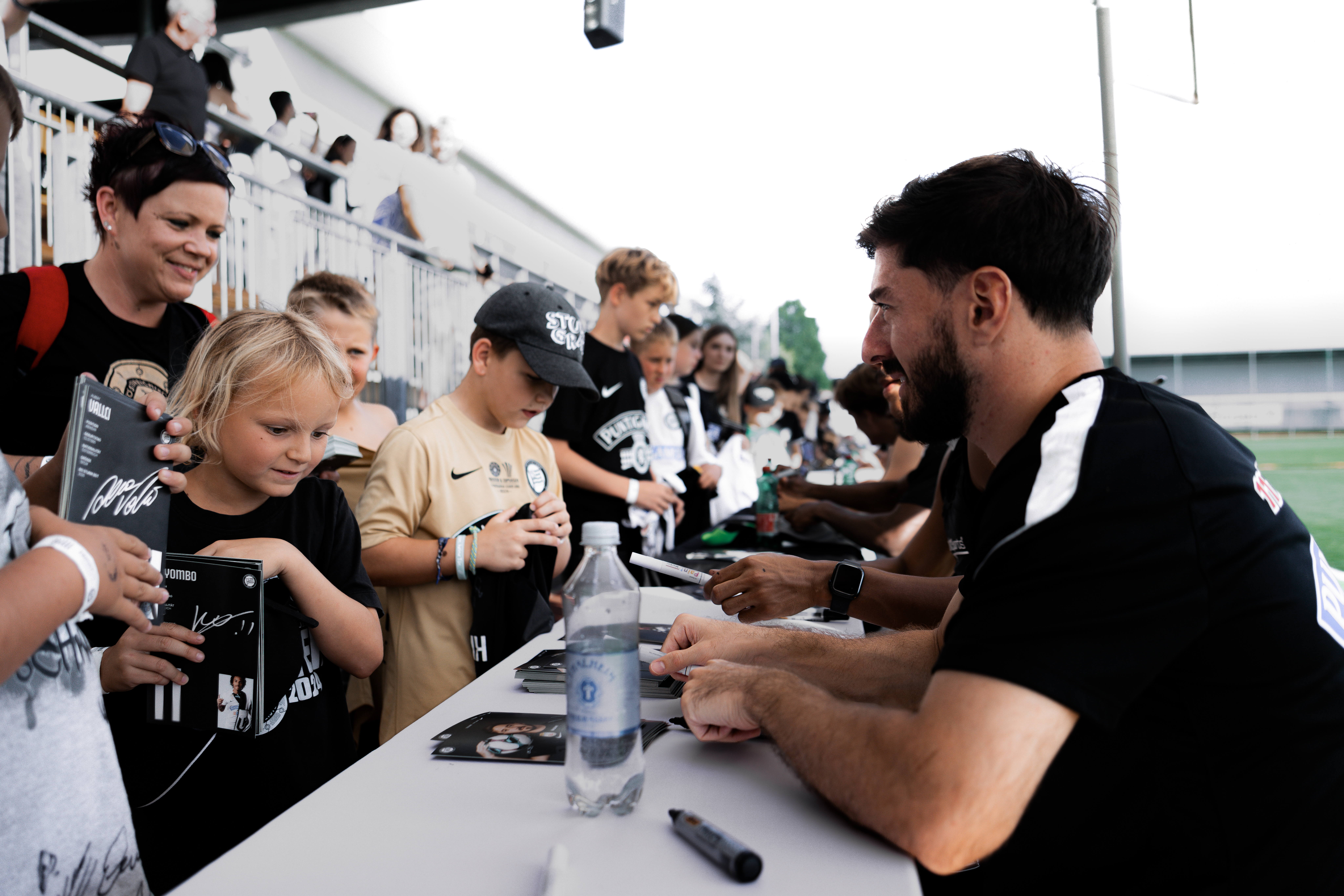 Kinder stehen vor einem Tisch und holen sich Autogramme von einem Fußballprofi