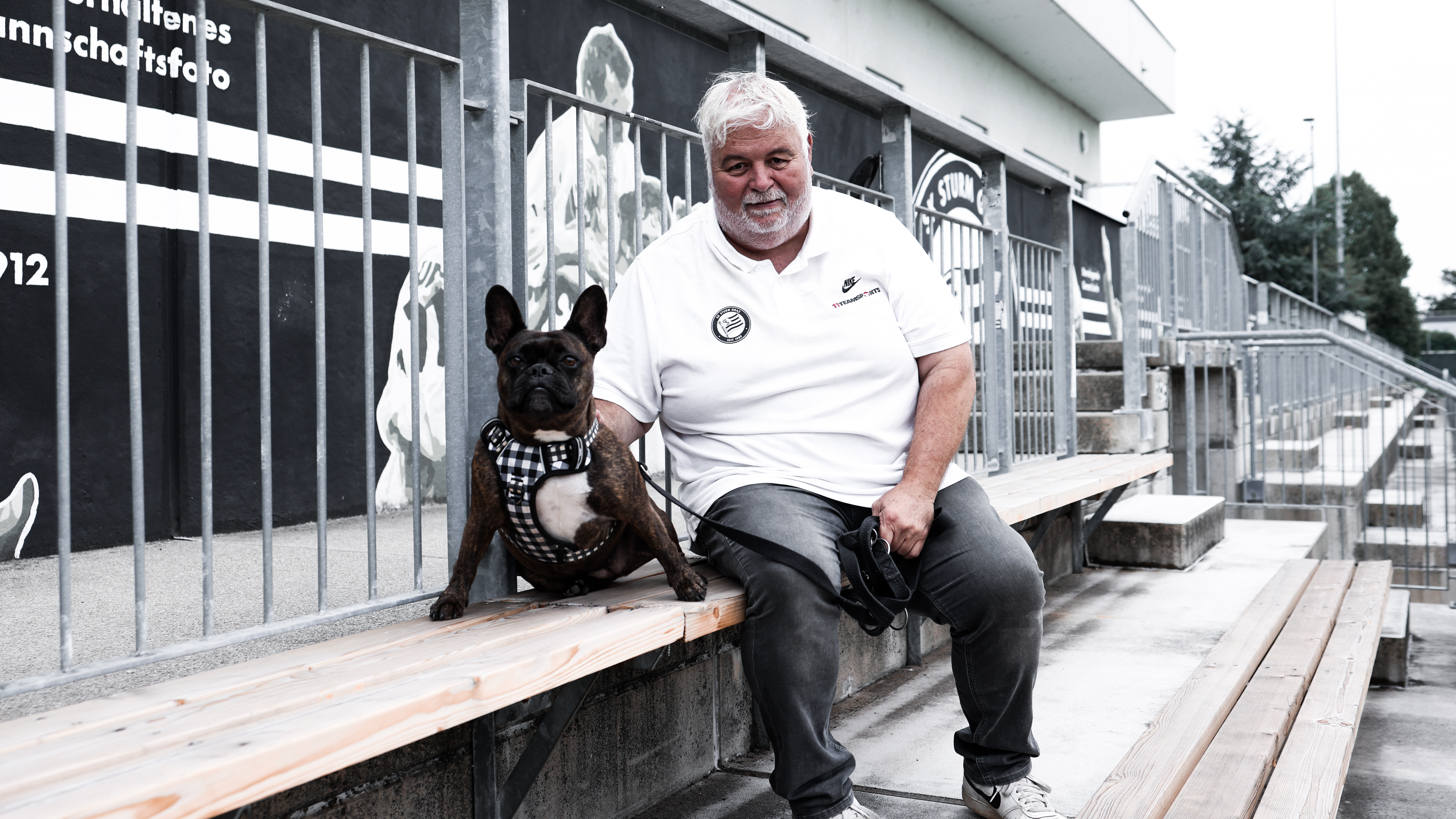 Ein Mann in einem weißen Shirt sitzt auf einer Holzbank und neben ihm sitzt ein Hund