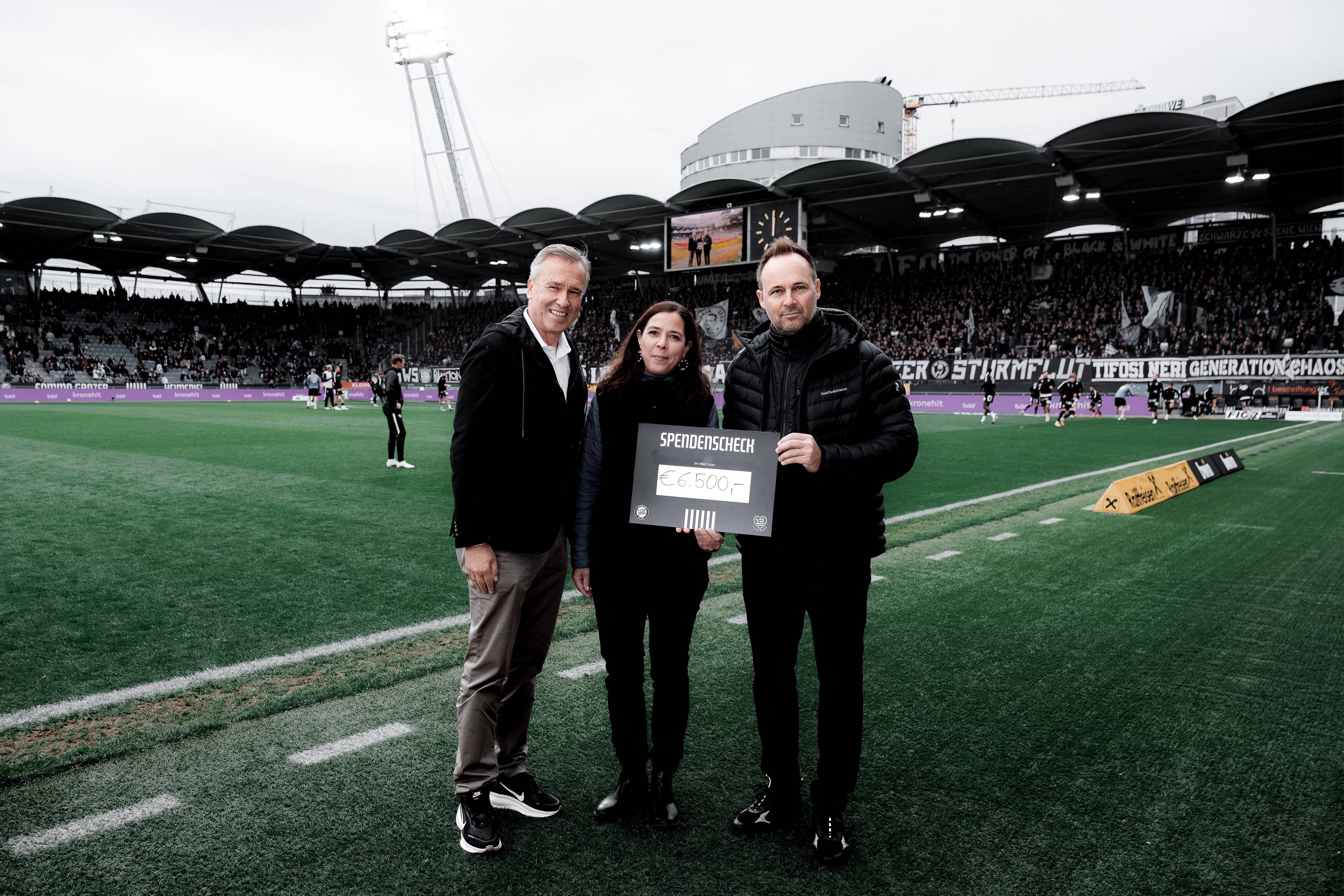Zwei Männer und eine Frau halten gemeinsam in einem gut besuchten Stadion einen Karton mit einem Aufdruck.