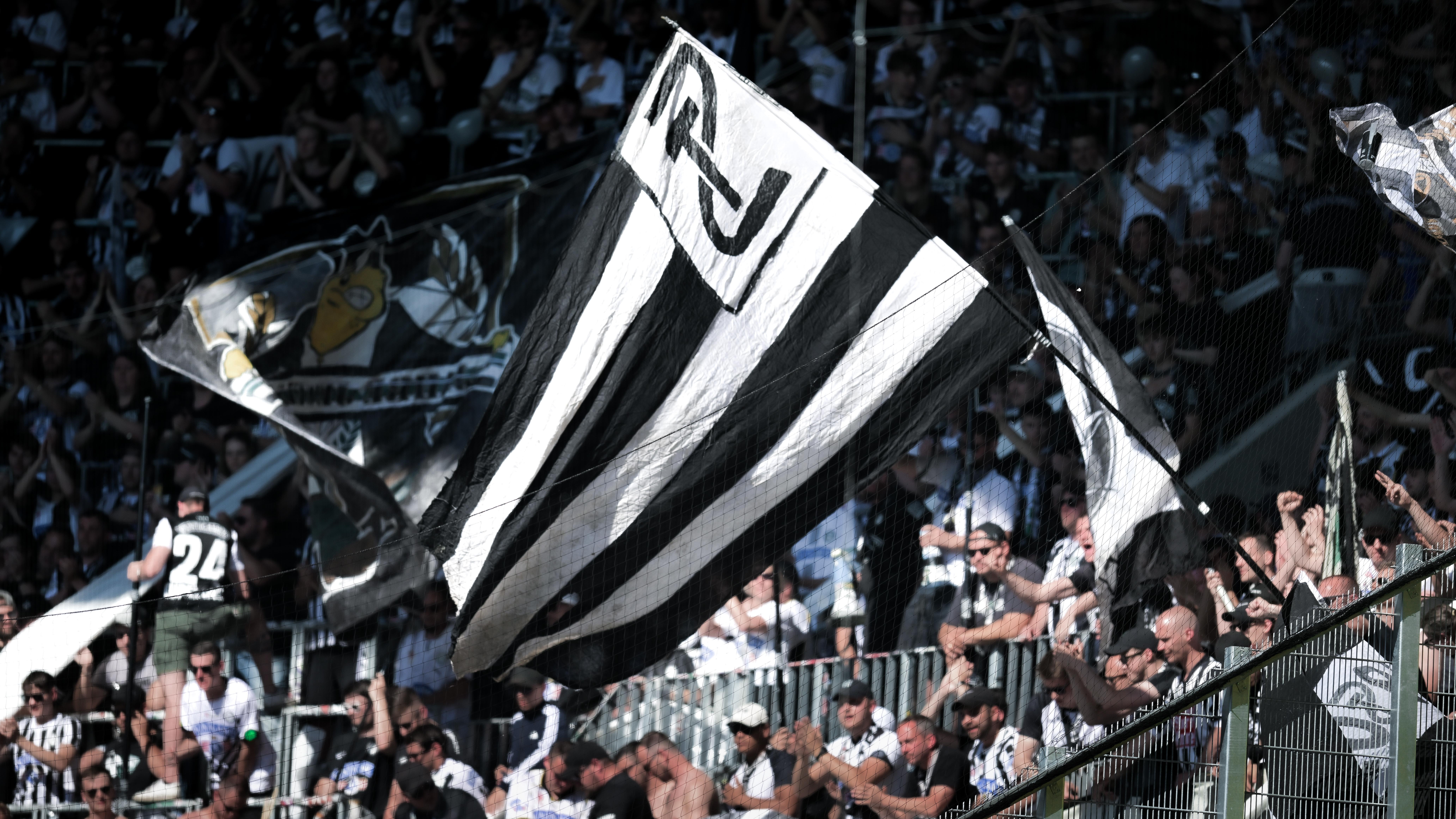 Eine große schwarz-weiße Fahne und viele Menschen in einem Bereich eines Stadions