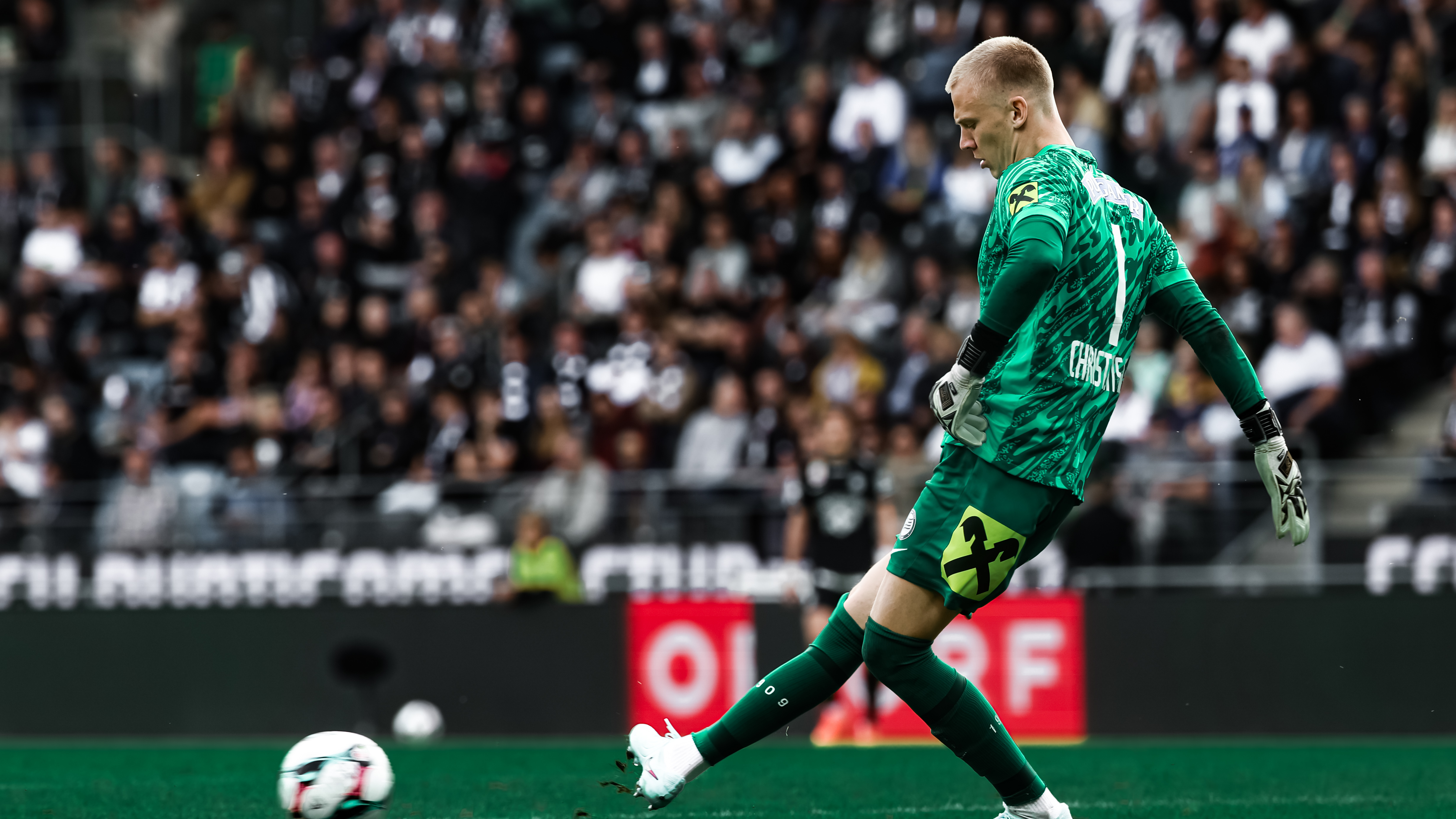 Ein Mann im grünen Trikot, der im Stadion mit vielen Fans einen Ball passt