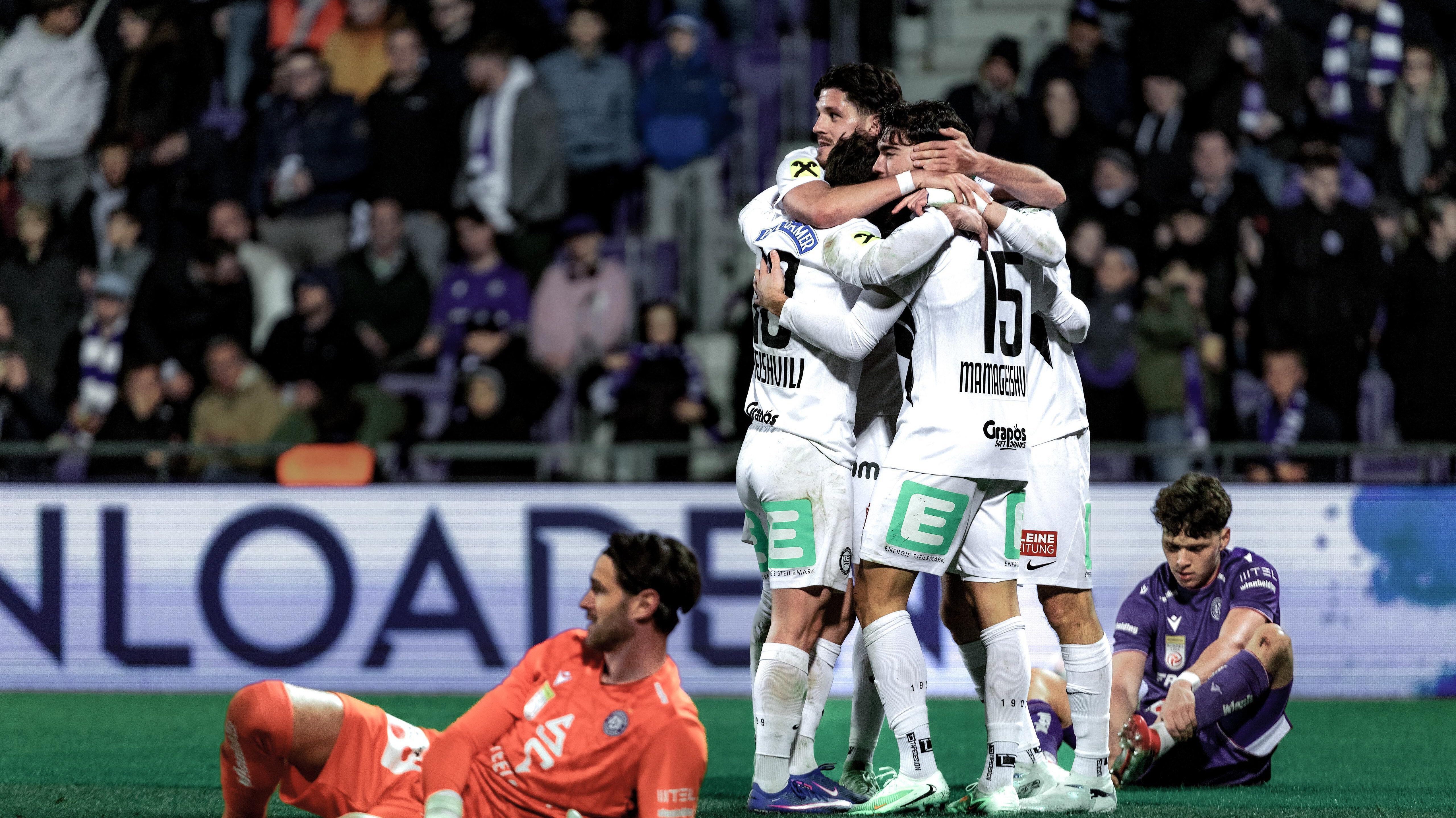 Vier Spieler in weißen Trikots umarmen sich, ein Spieler in violett und in Orange liegen am Boden.
