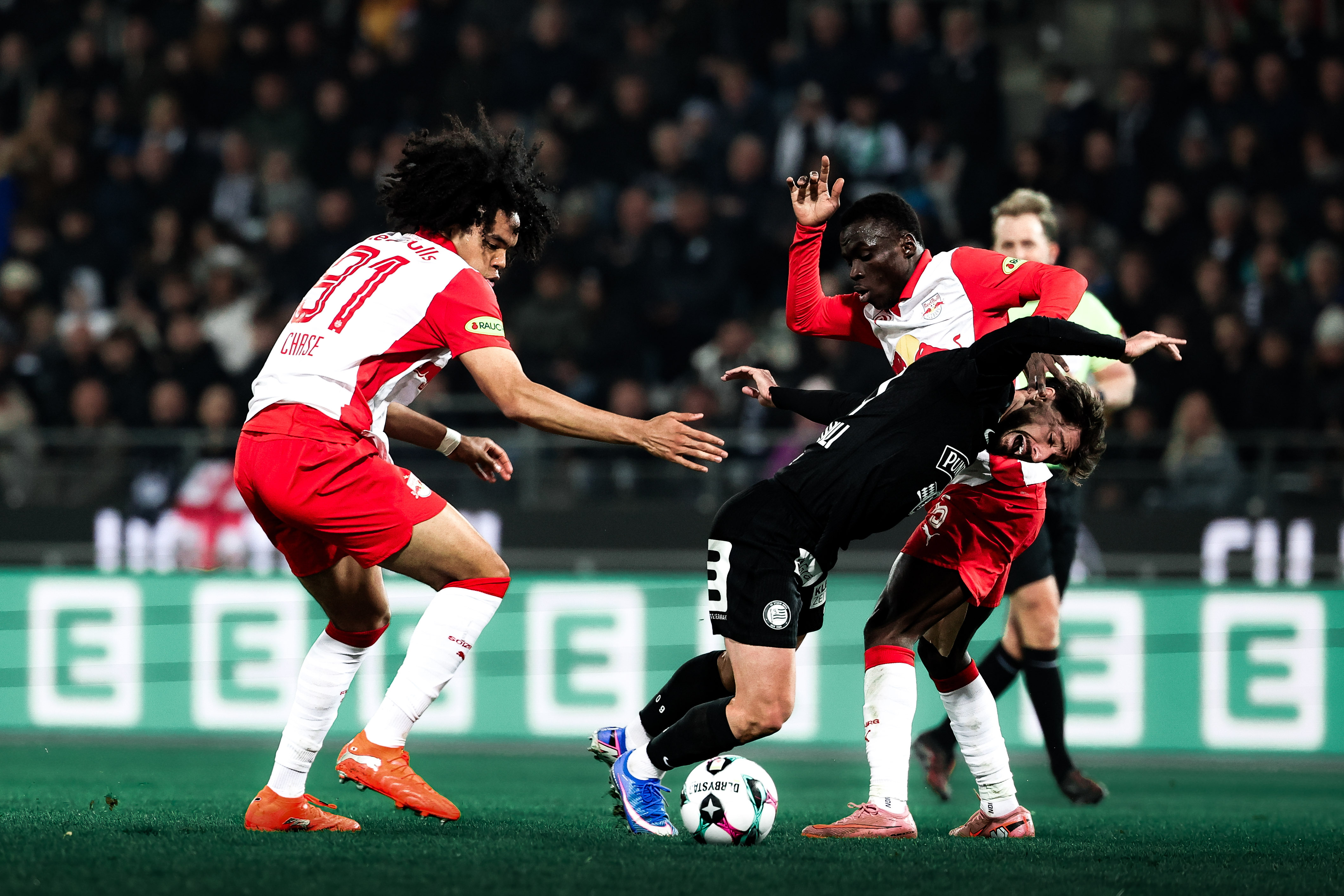 Zwei Spieler in rot und weiß kämpfen gegen einen Spieler in schwarz um einen weißen Ball.