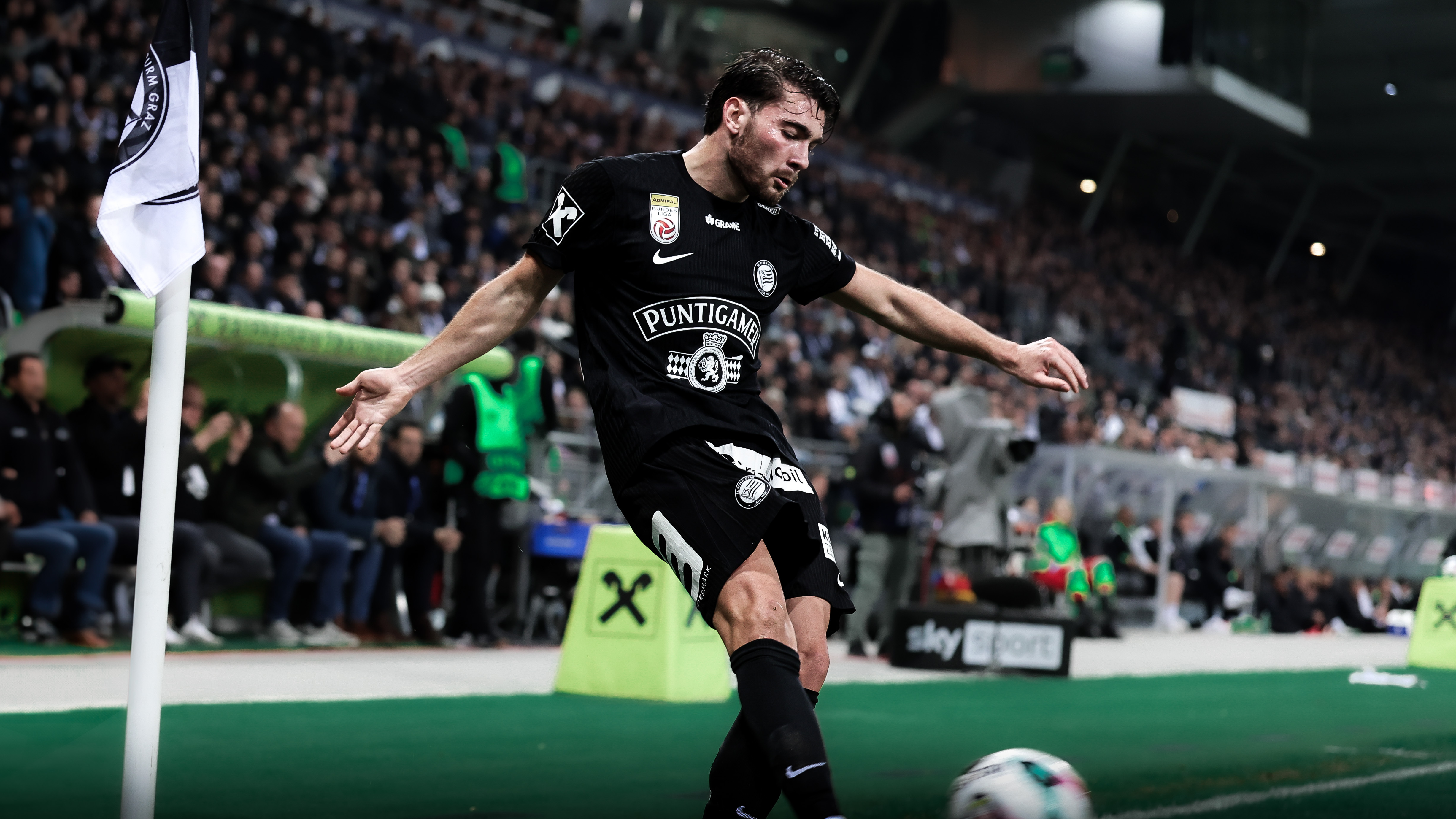 Ein Spieler in einem schwarzen Trikot und einer schwarzen Hose tritt gegen einen Ball in einem vollen Stadion im Hintergrund