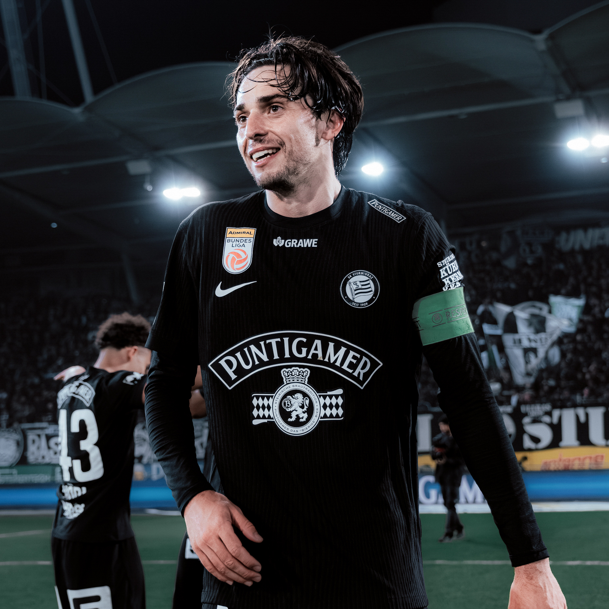 Ein Mann mit schwarzem Trikot und grüner Kapitänsbinde lächelt.