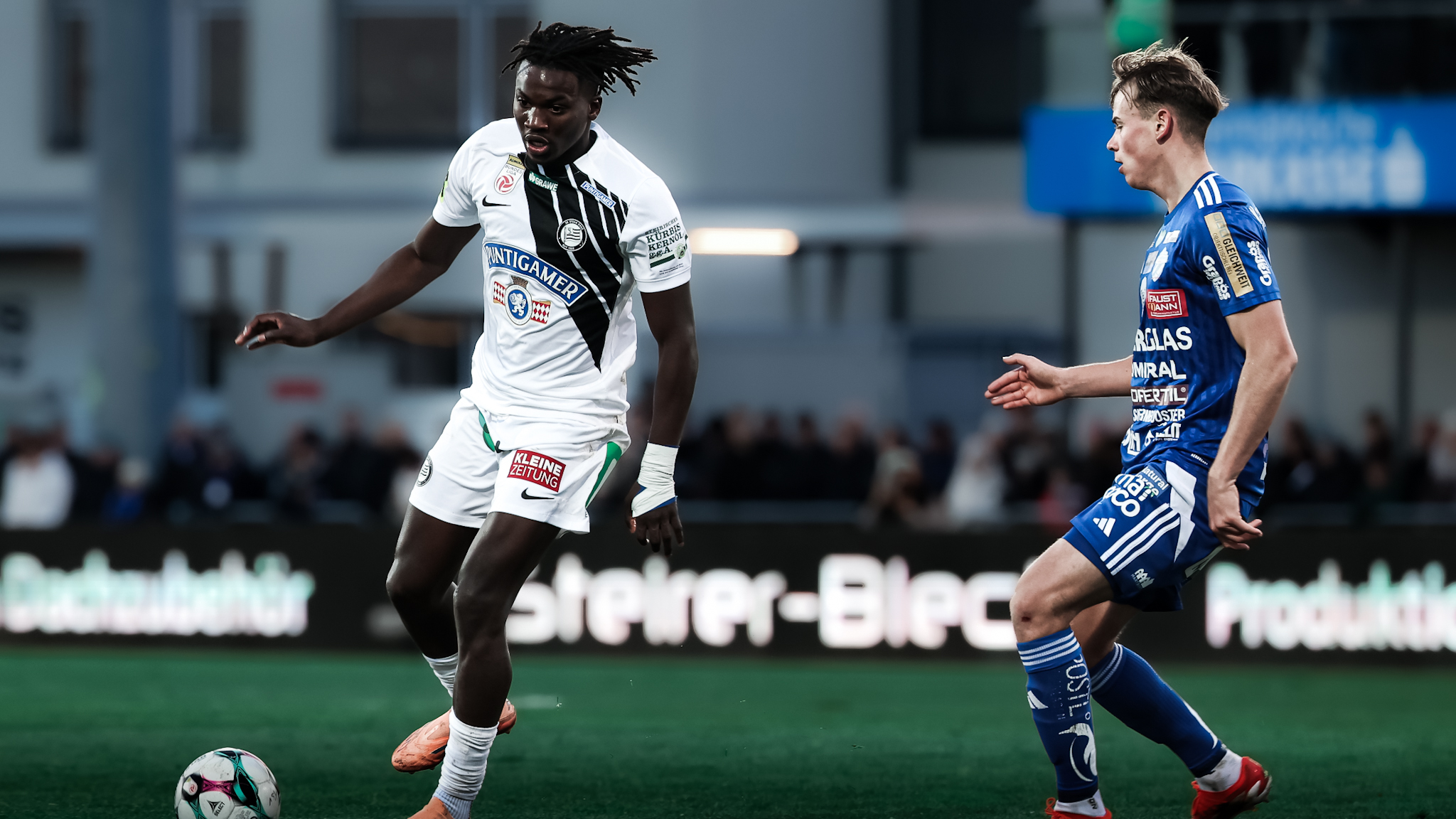 Ein Spieler in einem weißen Trikot hat den Ball und verteidigt ihn gegen einen Spieler in einem blauen Trikot