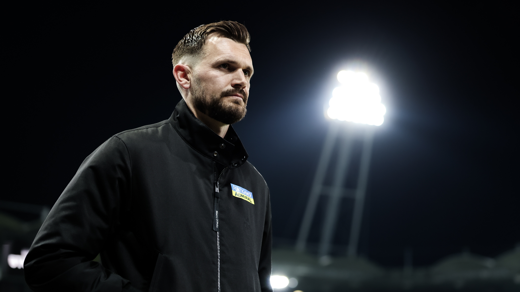 Ein Mann mit Bart und einer schwarzen Jacke geht in einem Stadion, im Hintergrund ist ein Flutlicht zu sehen.